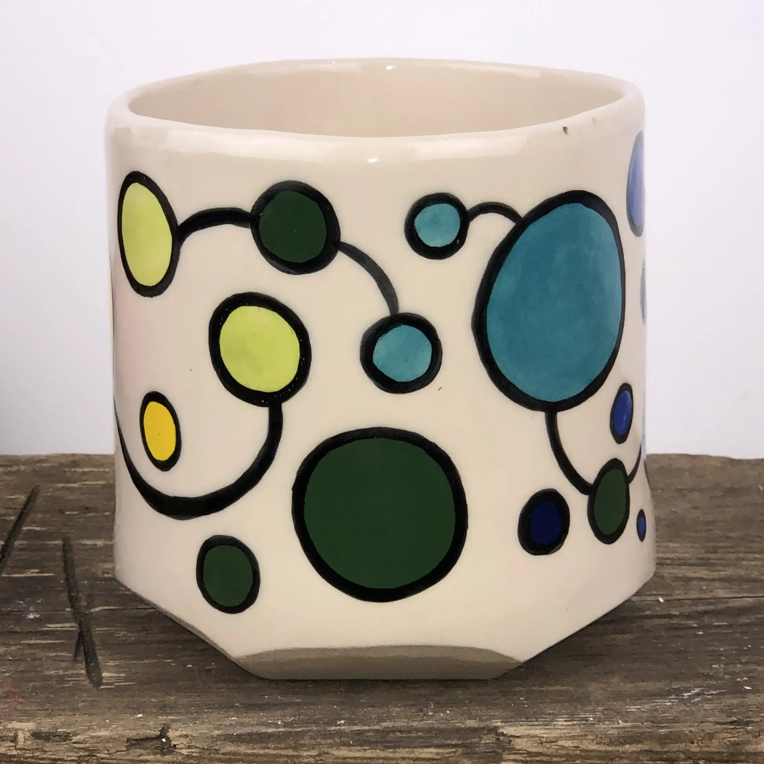 A ceramic planter with colourful circle patterns, featuring pale green, dark green and turquoise circles outlined in black, placed on a wooden surface.