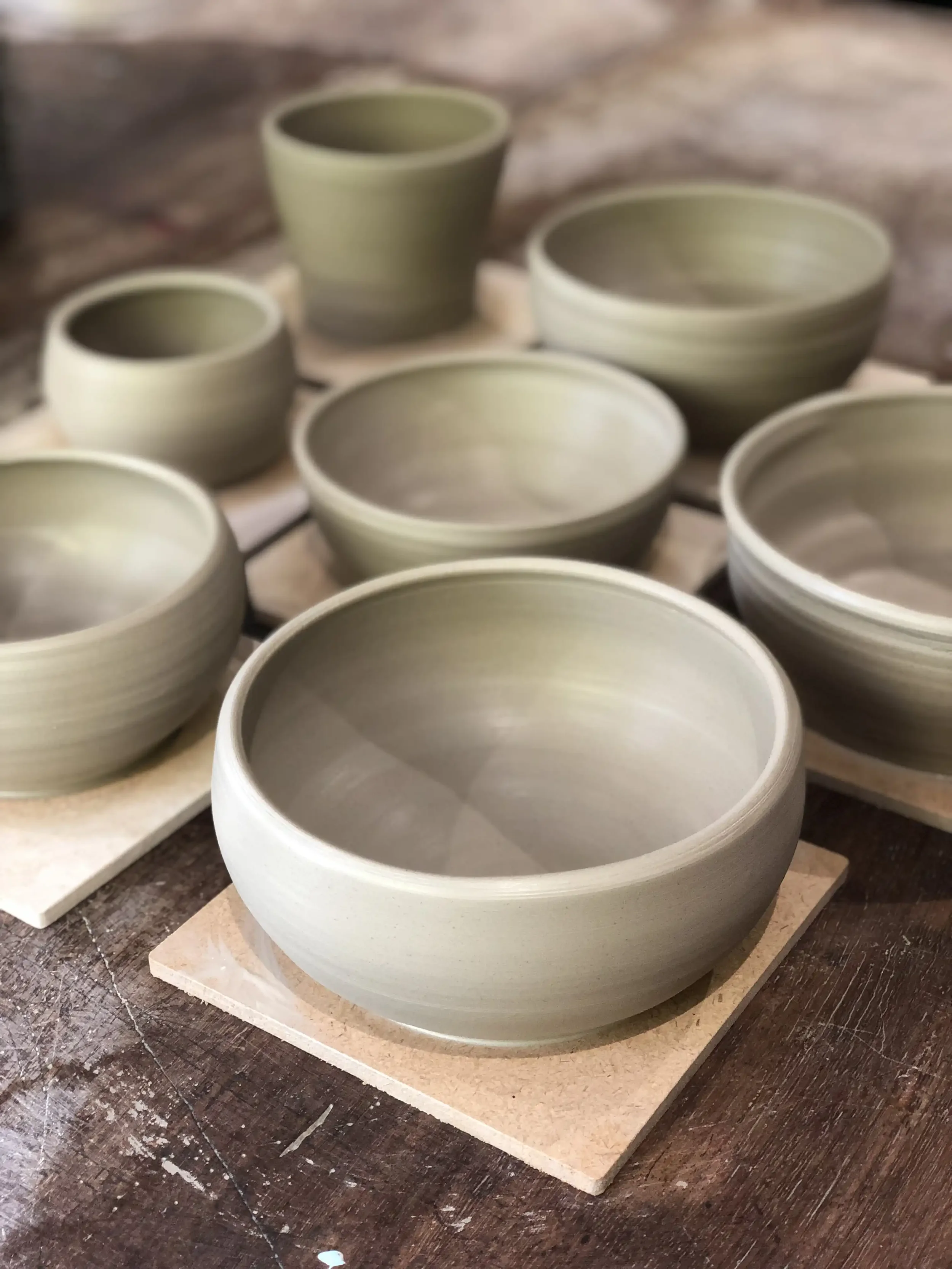 Several unglazed pottery bowls and cups on small tiles, arranged on a wooden table, in a pottery studio.