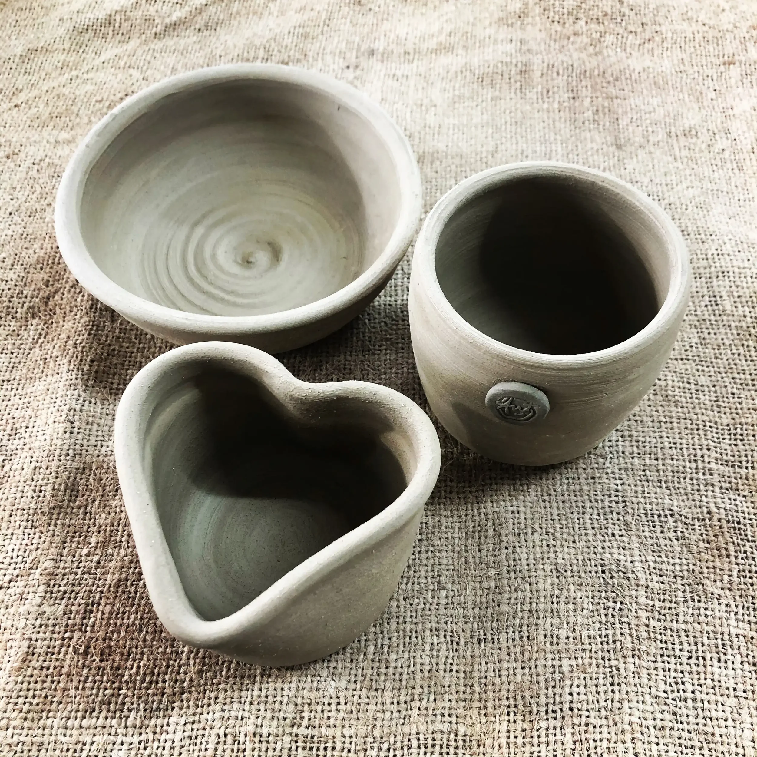 Set of three unglazed ceramic bowls of different sizes and shapes on a hessian surface, one small round, one larger round, and one heart-shaped.