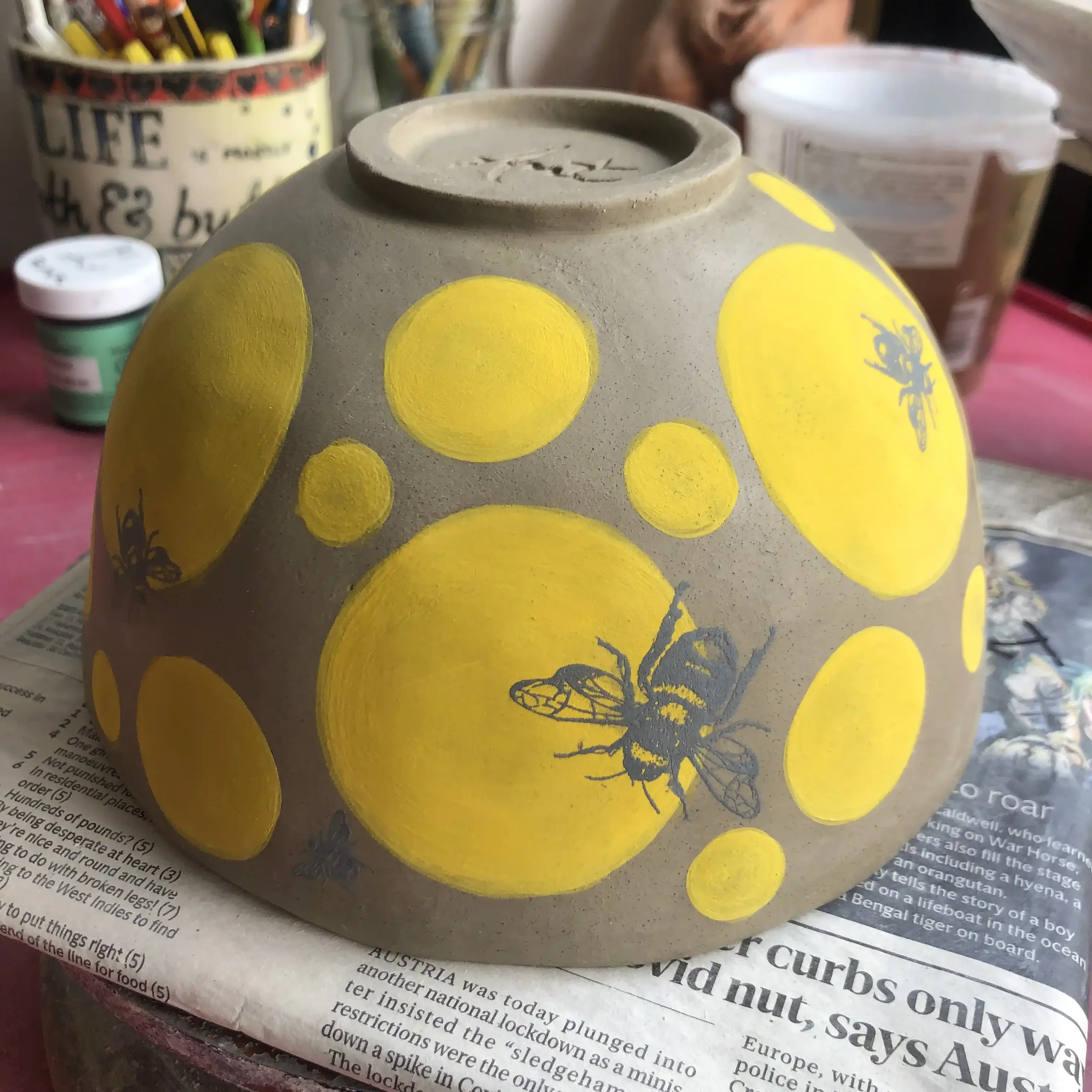 Hand-painted ceramic bowl decorated with yellow circles and black bee illustrations, placed on a newspaper surface with art supplies in the background.