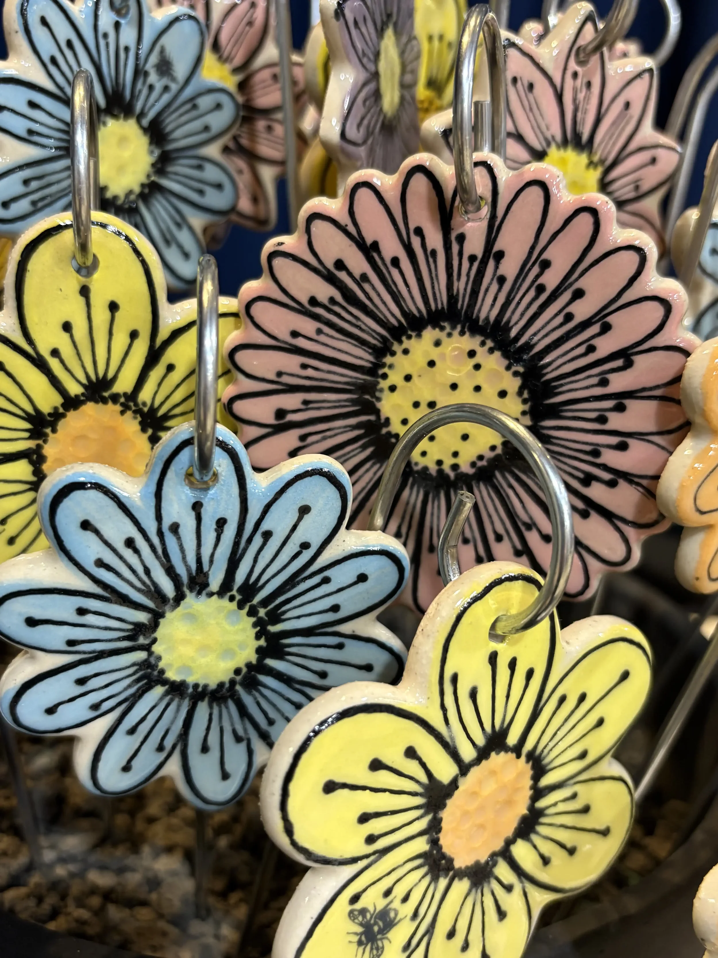 Colourful flower-shaped clay hanging decorations with black outlining and displayed on metal stakes.