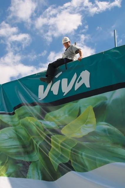 A man in a hard hat sitting on top of the 'viva' section of the viva Italia Safeway banner under a partly cloudy sky.