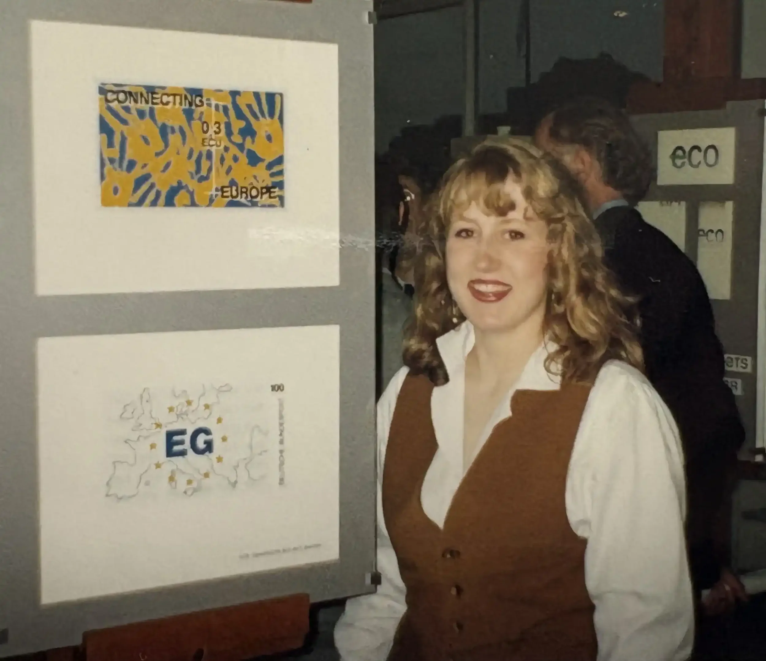 A woman with curly blonde hair and red lipstick smiling at an art exhibition, standing next to two framed posters on a grey wall. One poster shows an abstract yellow and blue stamp design.