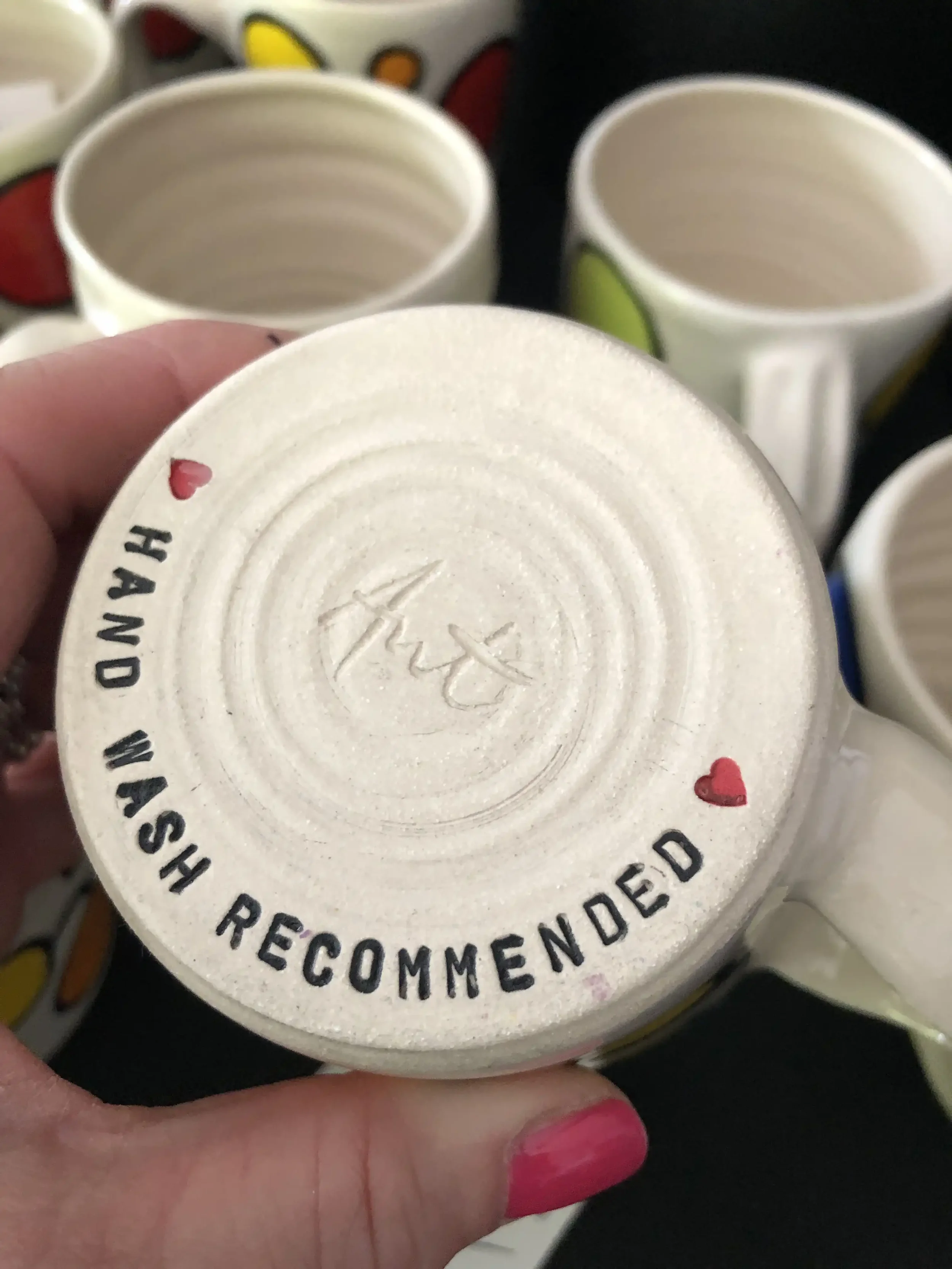 Bottom of a ceramic mug with the phrase 'HAND WASH RECOMMENDED' and small red hearts, surrounded by other colourful mugs with spot designs.