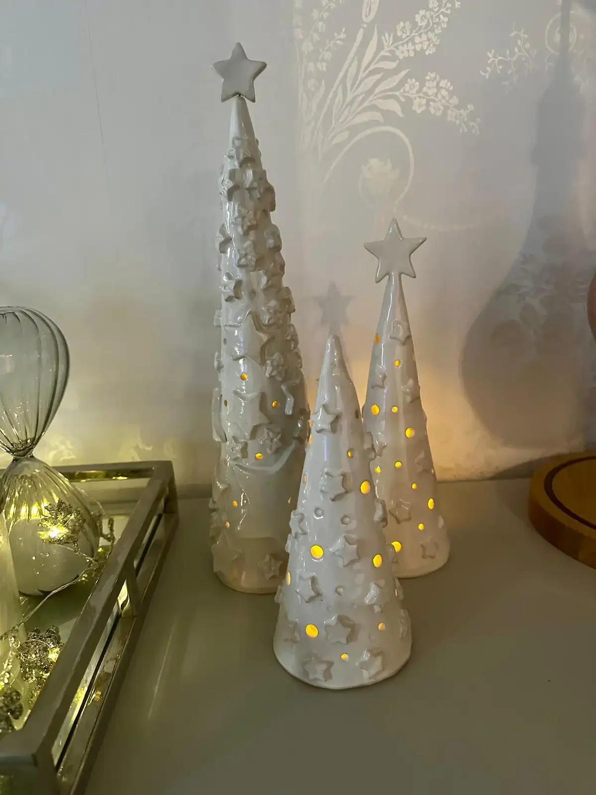Decorative white ceramic Christmas trees with star toppers and cut-out stars, illuminated from inside, on a table next to a glass display box with holiday ornaments.