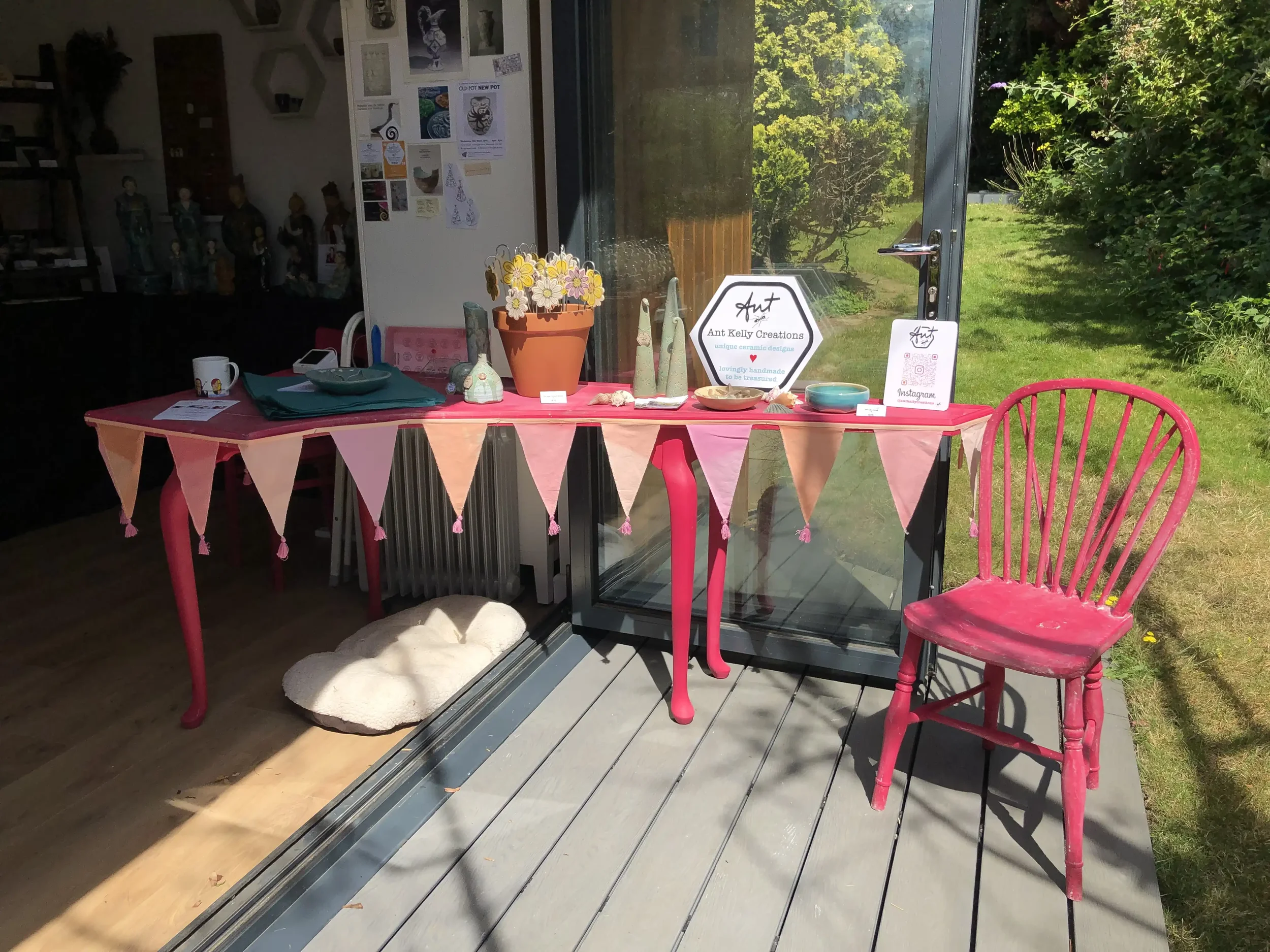 Pink table decorated with pink bunting, displaying ceramic art pieces and a sign for Ant Kelly Creations open studio event, situated on a deck outside Ant's garden studio with bi-fold doors, overlooking the grass and trees in the garden.