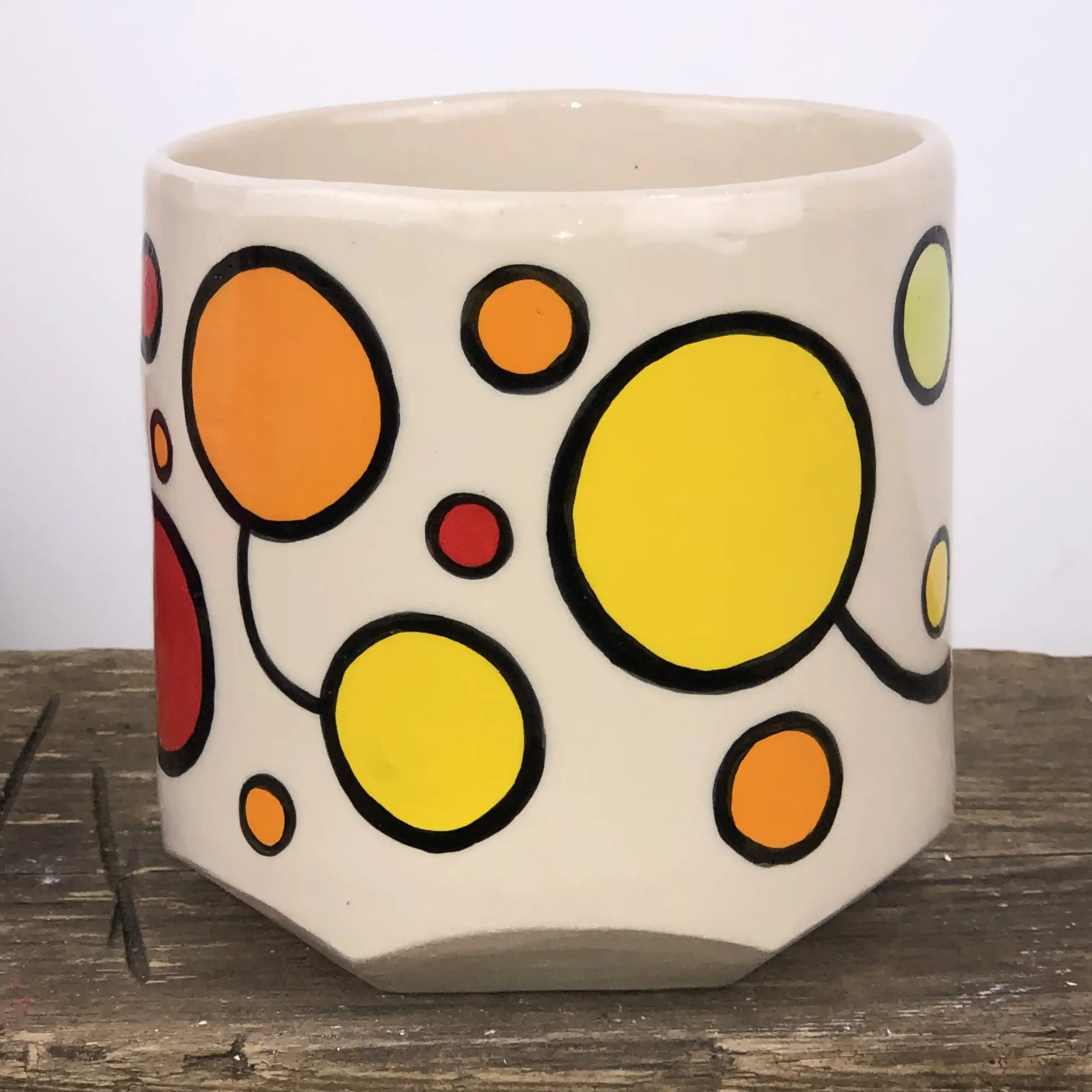 A ceramic planter with colourful circle patterns, featuring yellow, orange, red, and pale green circles outlined in black, placed on a wooden surface.