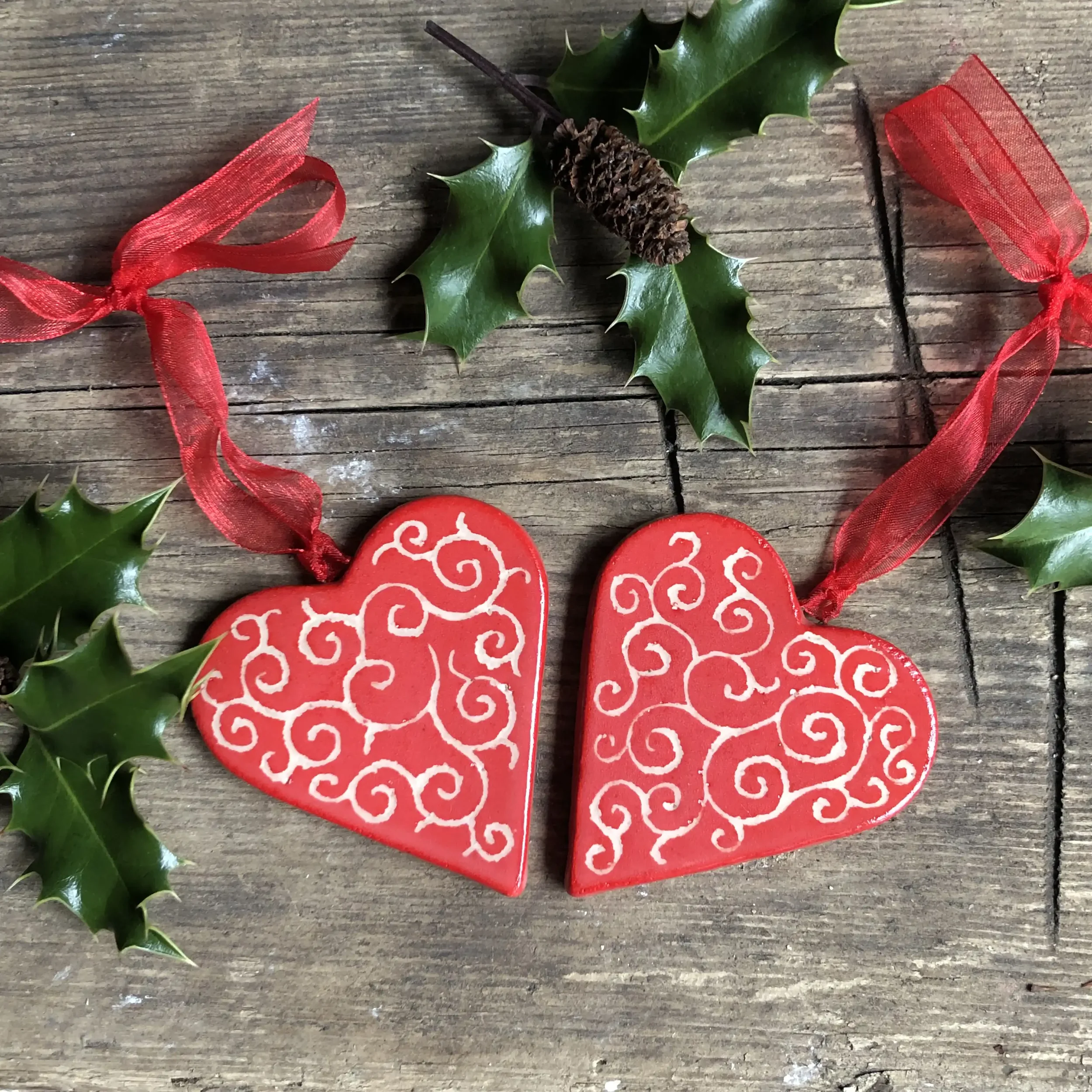 Two red heart-shaped ornaments with white swirling patterns, attached to red ribbons, surrounded by holly leaves and pine cone on a wooden surface.