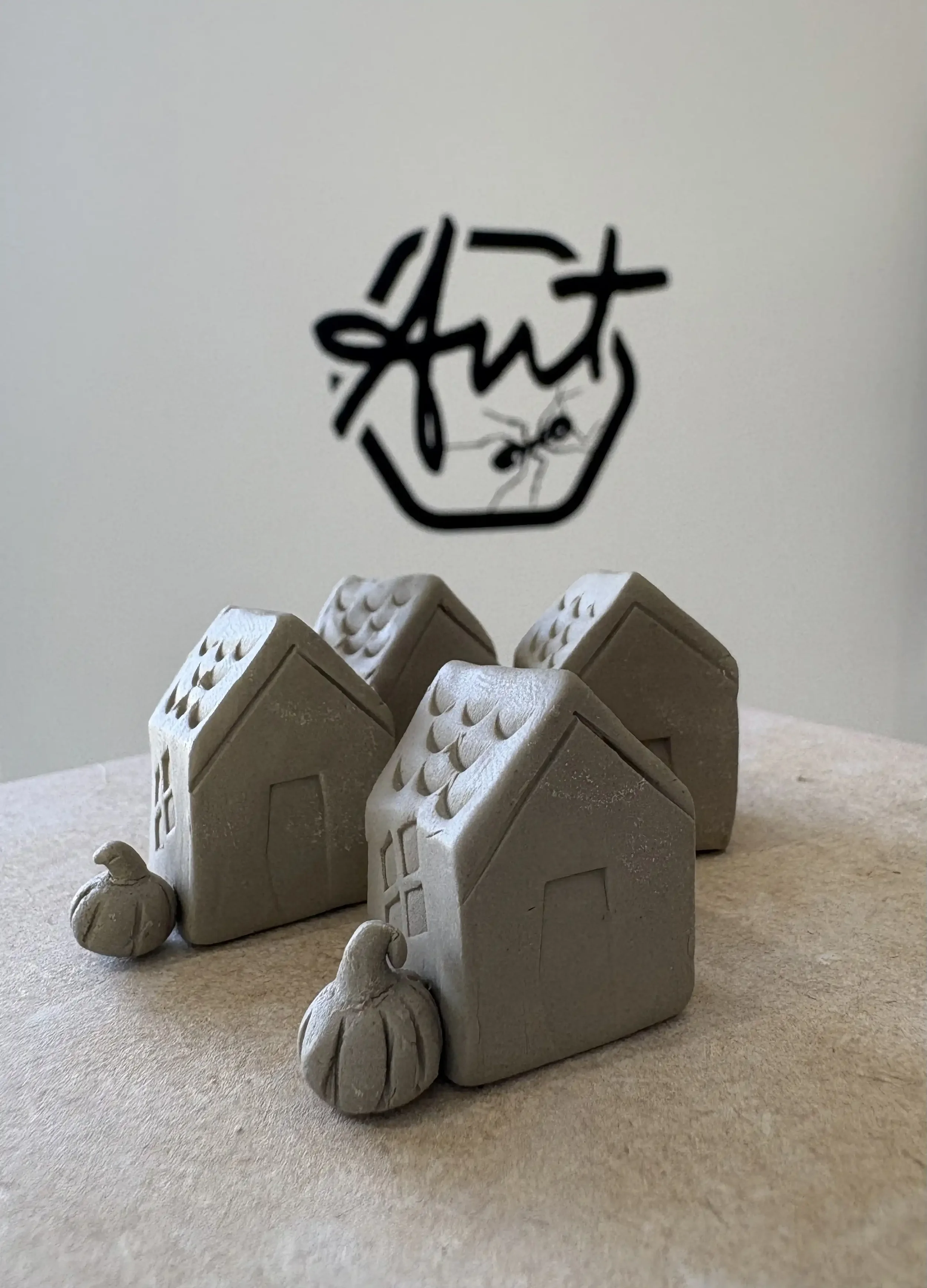 Three small ceramic house figurines with pumpkins in front, placed on a beige surface, with a white wall behind them featuring the Ant Kelly Creations logo in black.