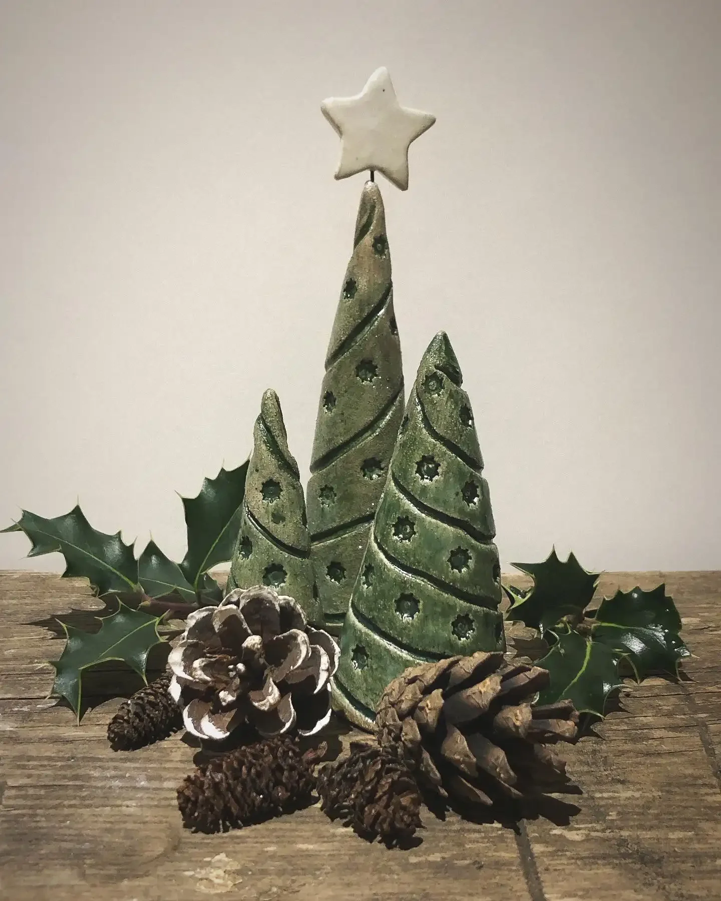 Decorative Christmas arrangement with three ceramic tree ornaments, pine cones and holly leaves on a wooden surface.