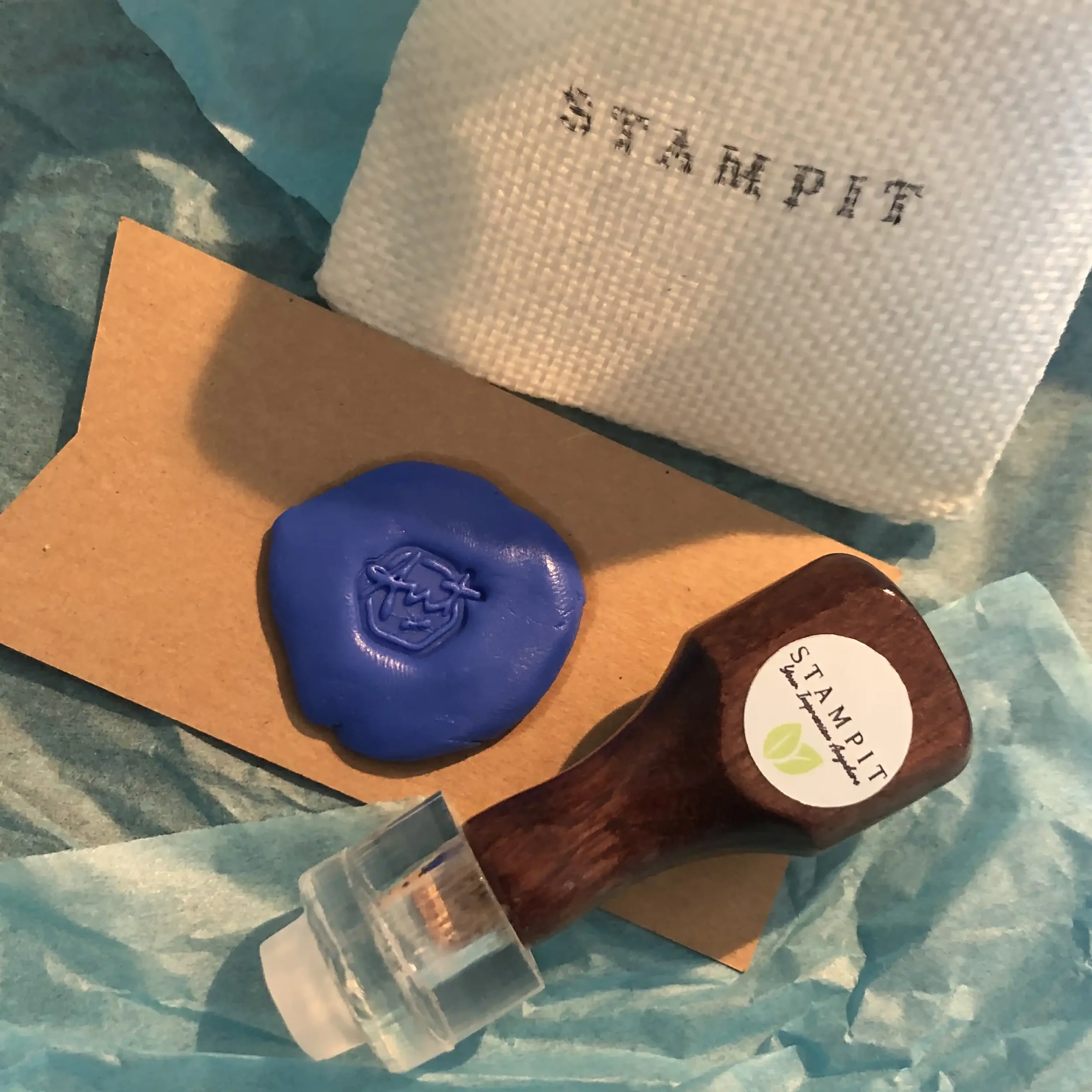 A sealing wax stamp with the label 'STAMPT IT' and a blue wax seal with an embossed logo, along with a white cloth bag with black text, all arranged on turquoise tissue paper.