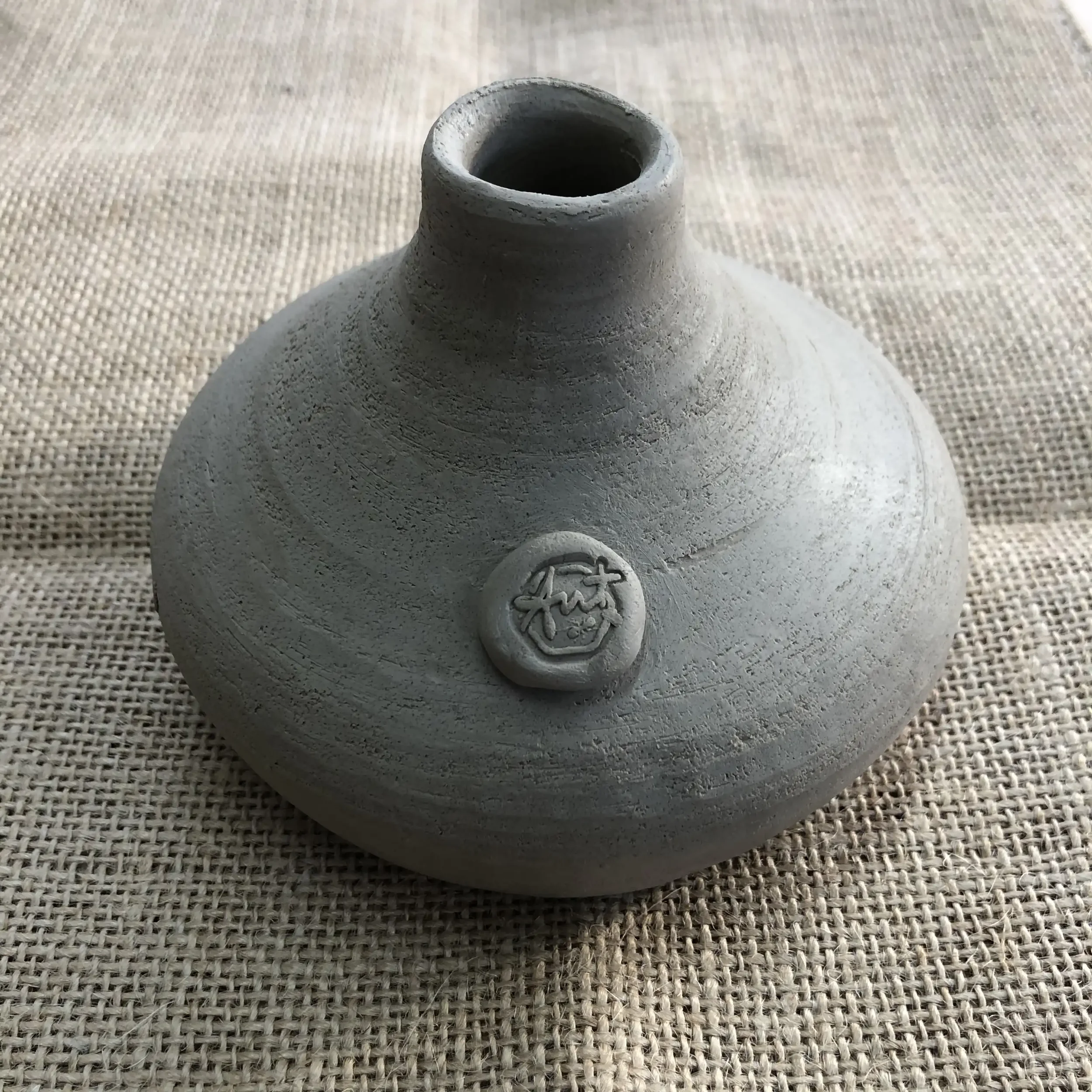 Unfired ceramic bud vase with a round body, narrow neck, and Ant Kelly Creations maker's mark button on the front, placed on a hessian surface.