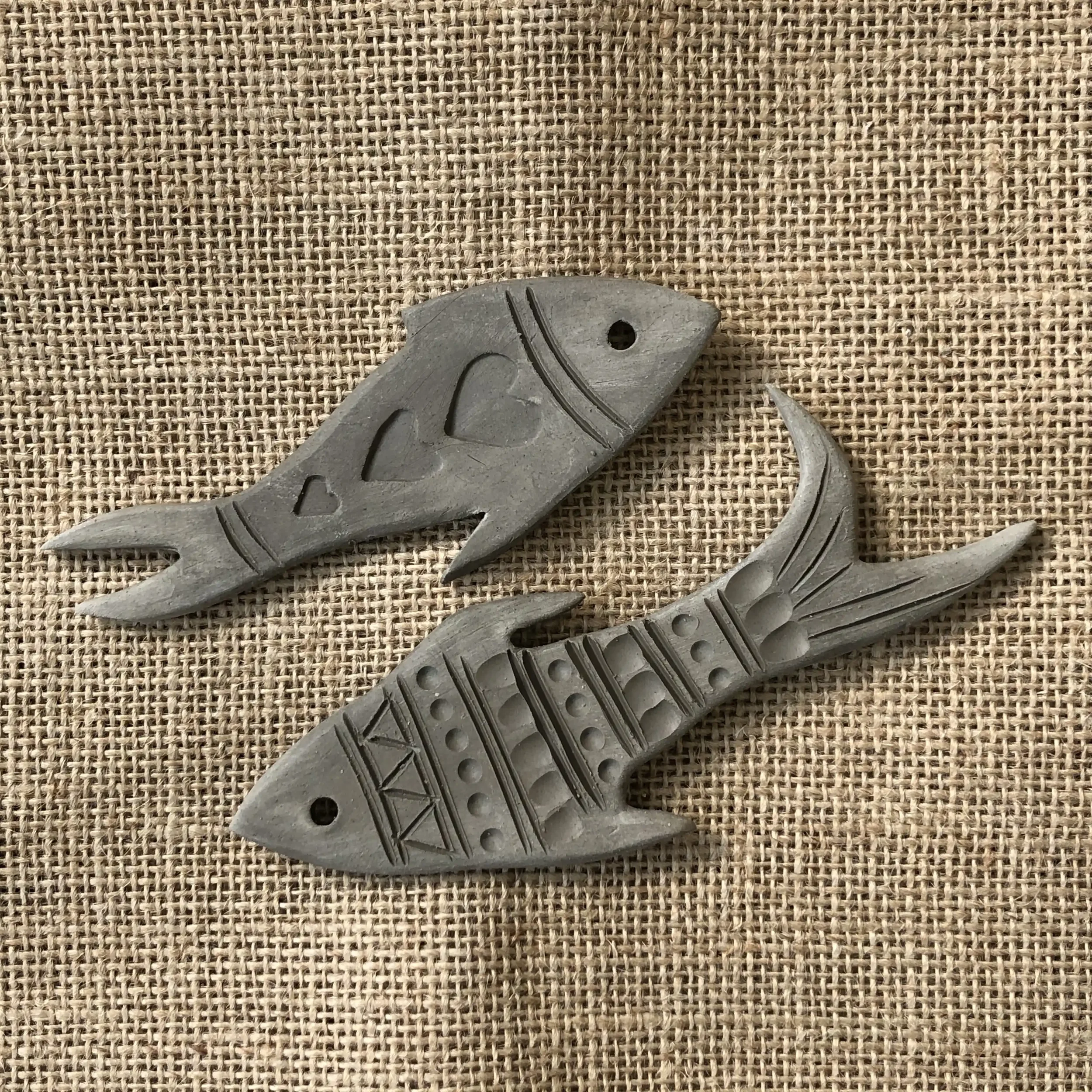 Two carved clay fish shapes with tribal patterns and cutouts, placed on a textured burlap fabric background.
