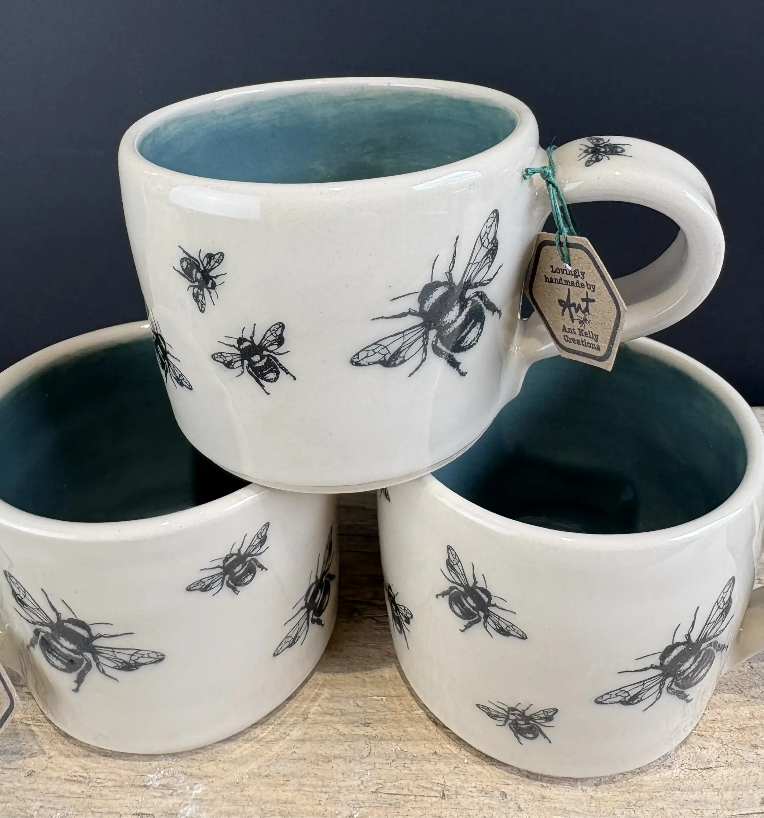 Set of three white ceramic mugs with black bee illustrations, one mug stacked on top of two others on a wooden surface.