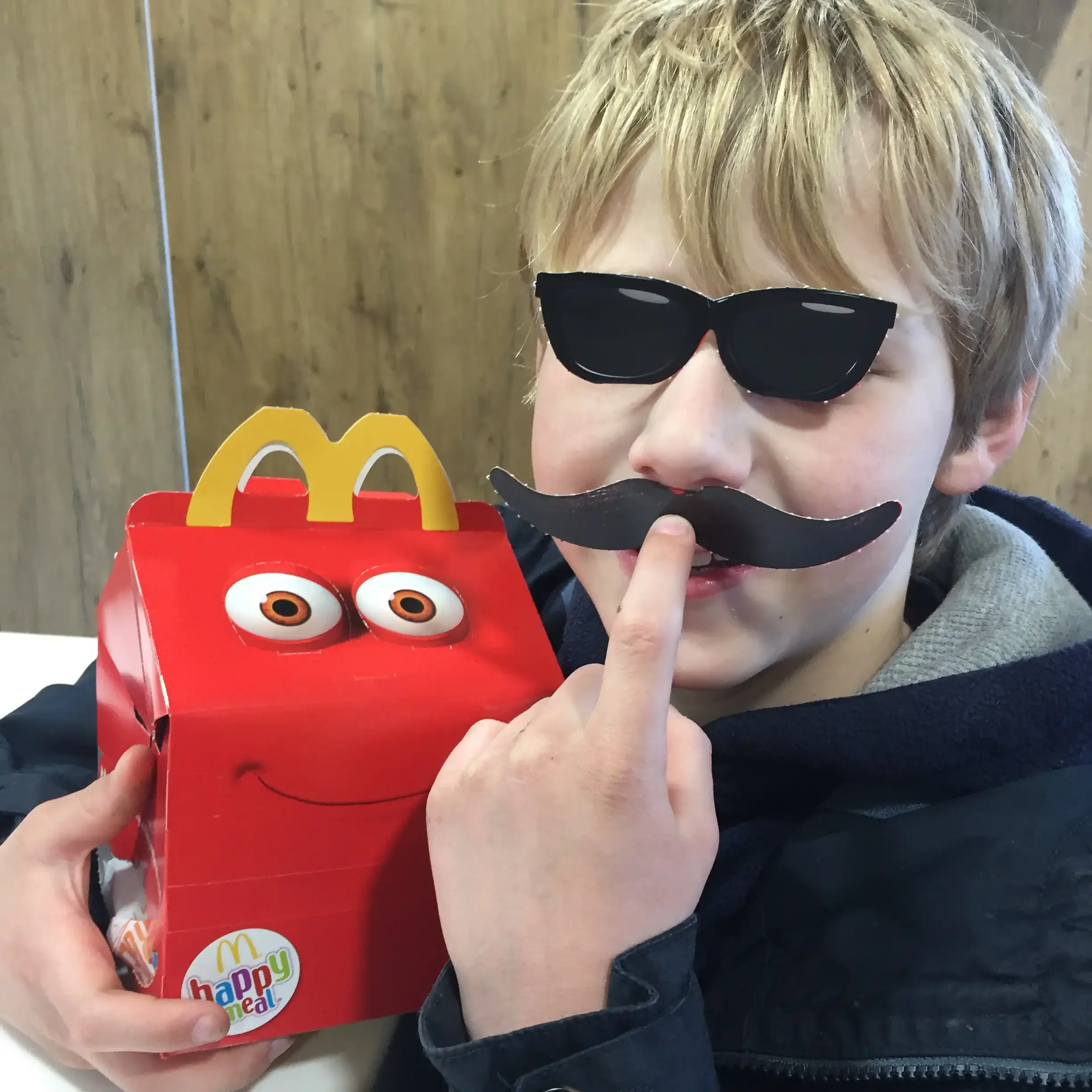 A boy with pop-out cardboard sunglasses and a fake moustache poses with a McDonald's Happy Meal box that has eyes and a smile.