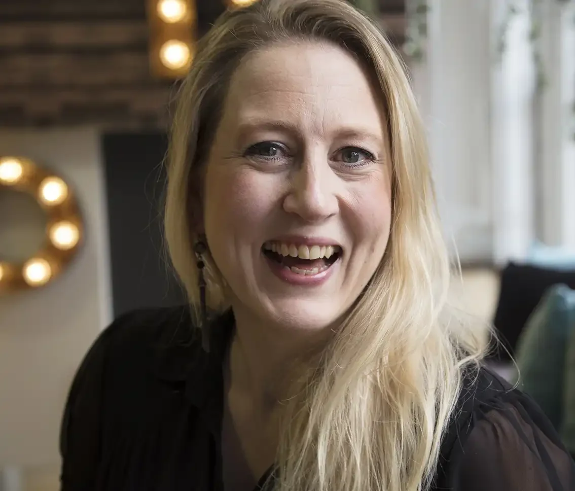 A smiling woman with long blonde hair, wearing a black top, in a cozy indoor setting with warm lighting and decorative elements in the background.