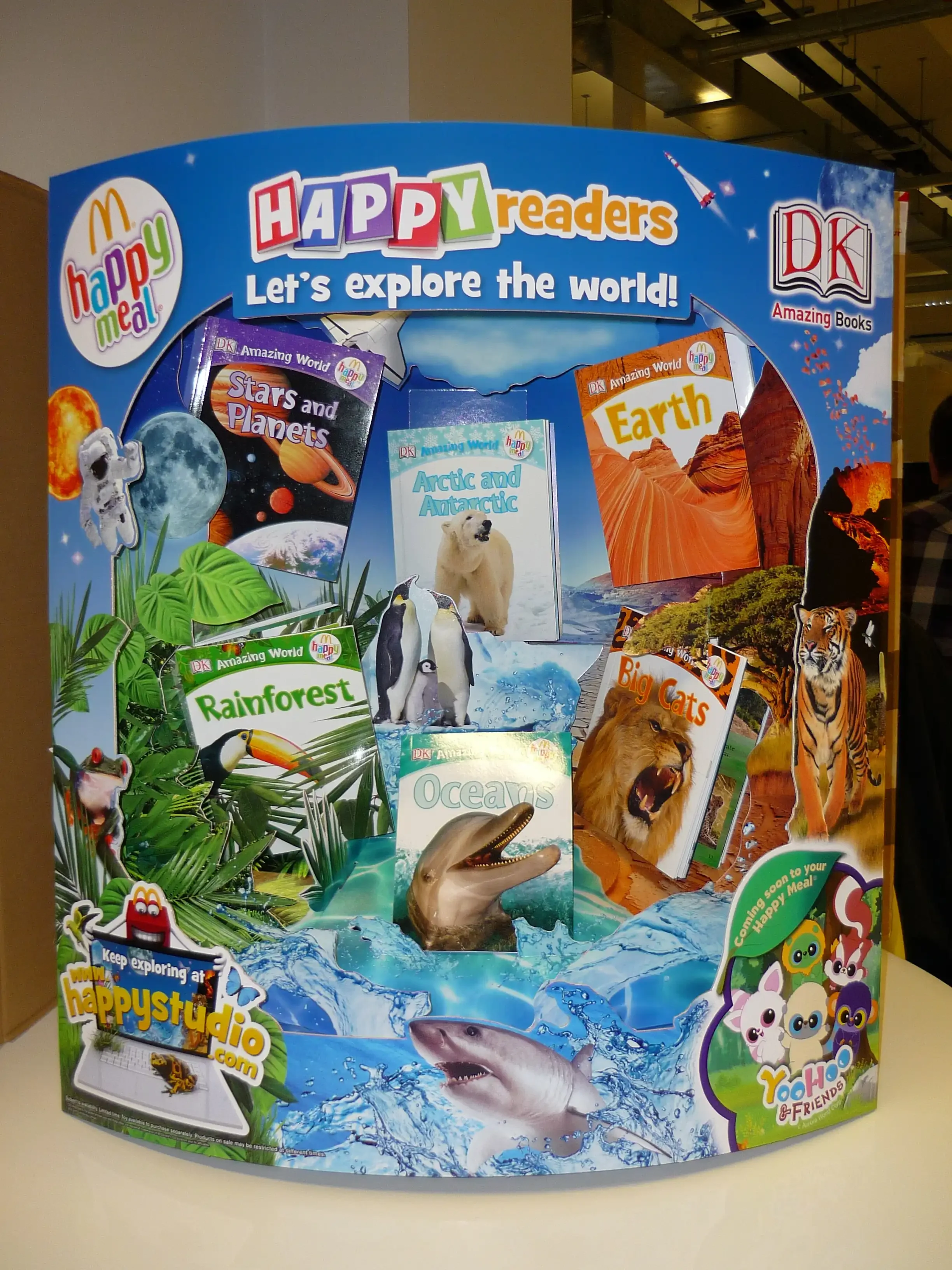Display of children's books titled "Stars and Planets," "Arctic and Antarctic," "Earth," "Rainforest," "Oceans," and "Big Cats" from the Happy Meal Happy Readers series, with an ocean scene with a shark, dolphin, penguins, and a polar bear.