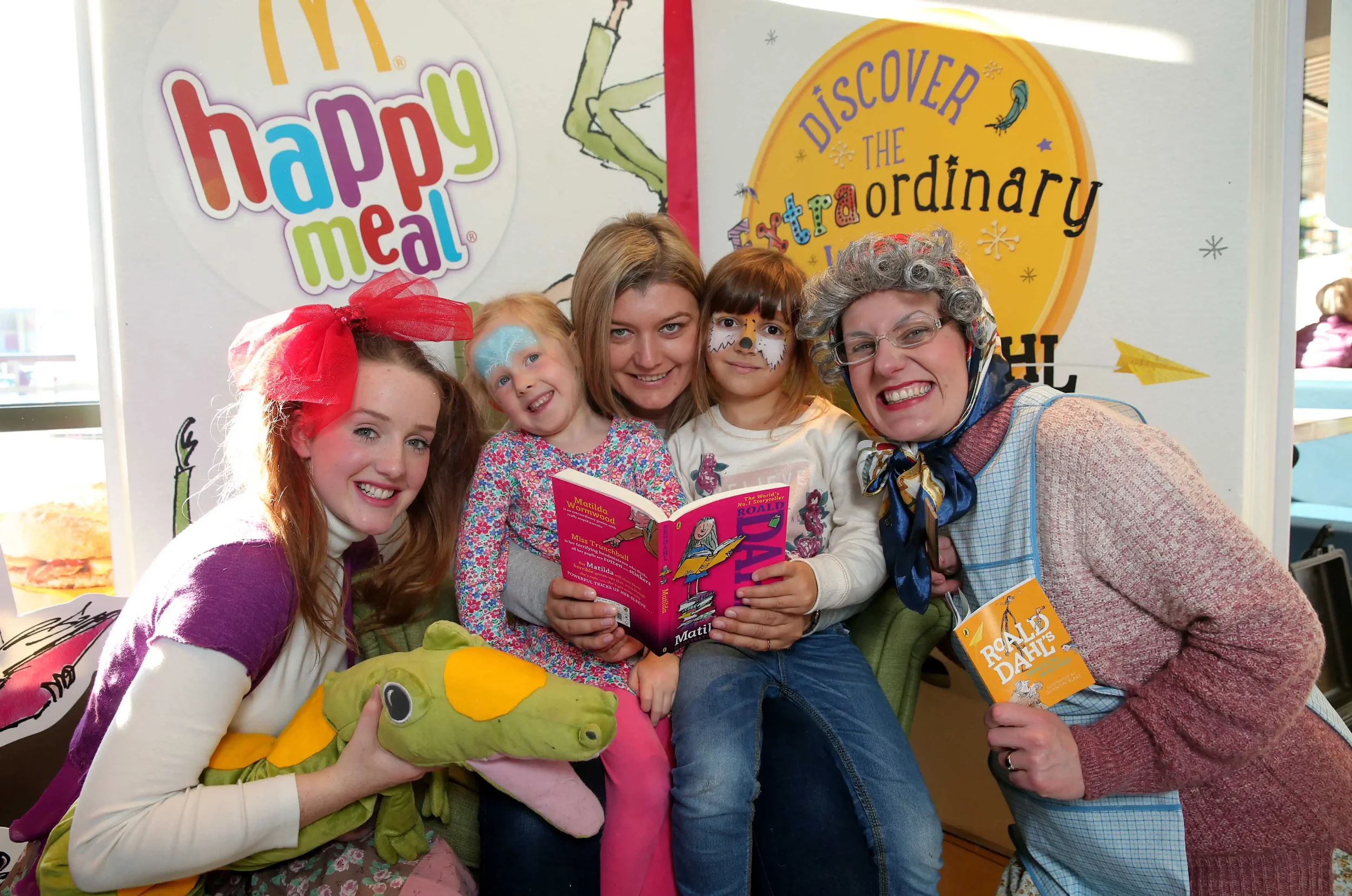 A group of five people, including children and an adult, smiling and sitting together. They are at an event or promotion for children's books, with colorful signage behind them. One girl is holding a soft green plush toy, and another girl has face pa