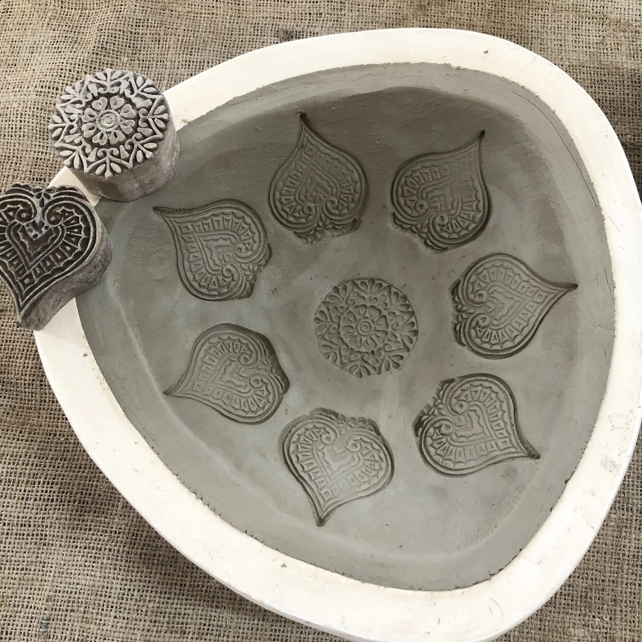 Wooden stamps with intricate patterns stamped onto a slab of clay inside a three-sided plaster mould on a hessian surface.