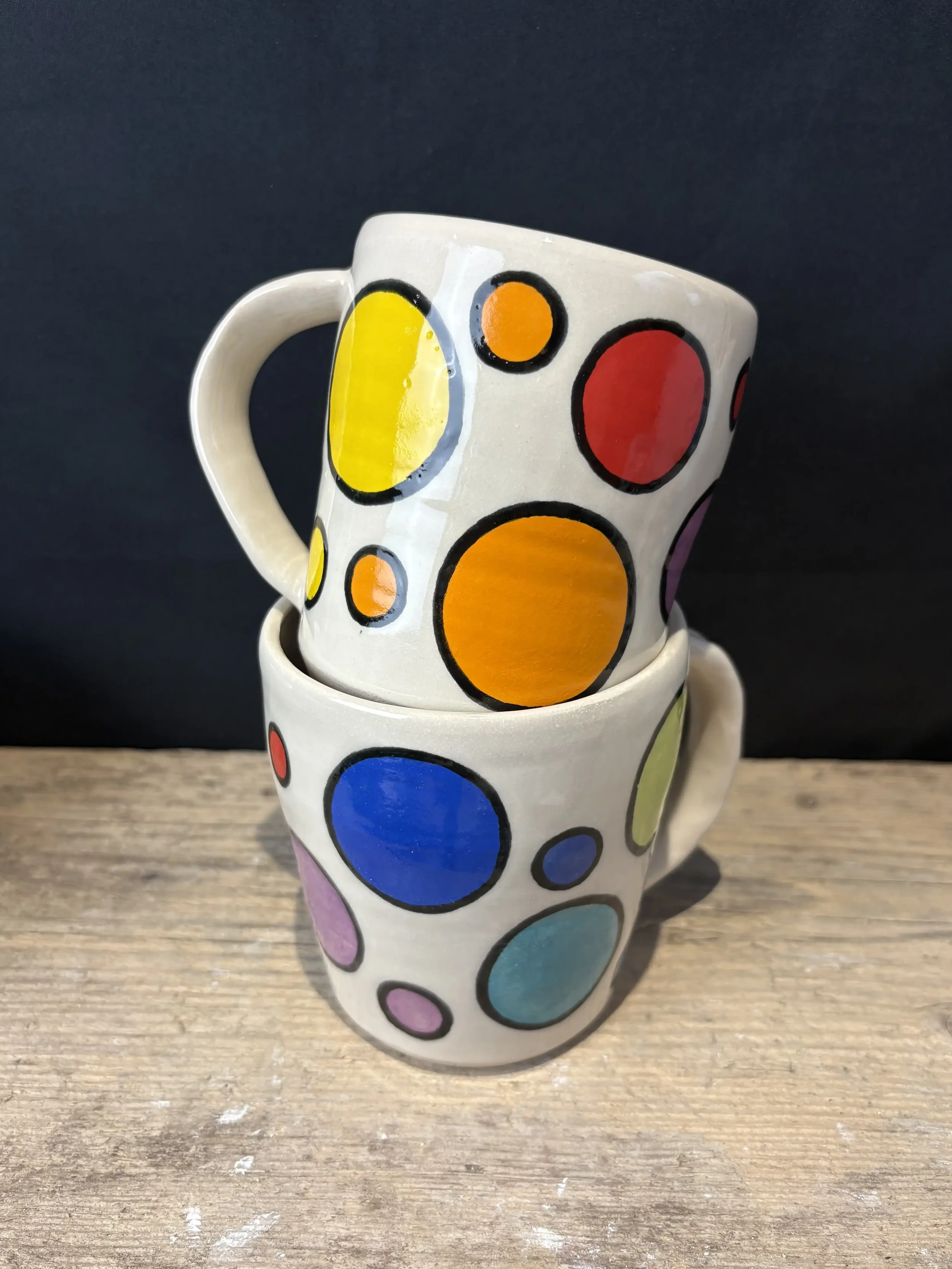 Two colourful ceramic mugs with abstract dot pattern in yellow, orange, green, blue, purple, and red, arranged on a wooden surface with a black background.