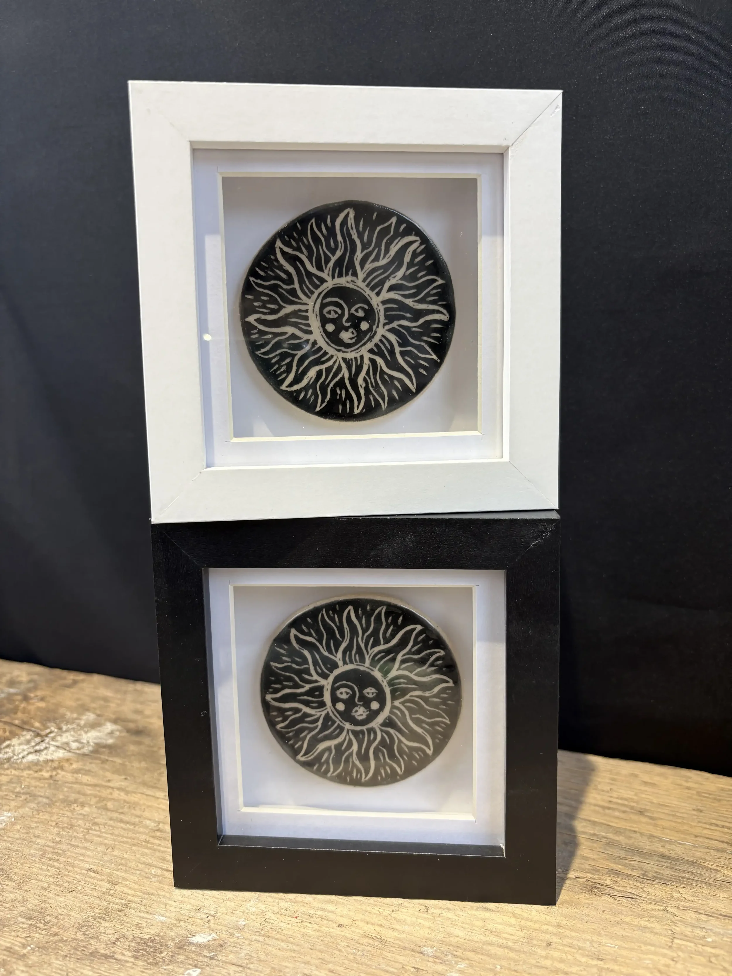 Two framed glazed tiles of a black circular design with a sun and moon face, one in a white frame and one in a black frame, placed on a wooden surface against a dark background.