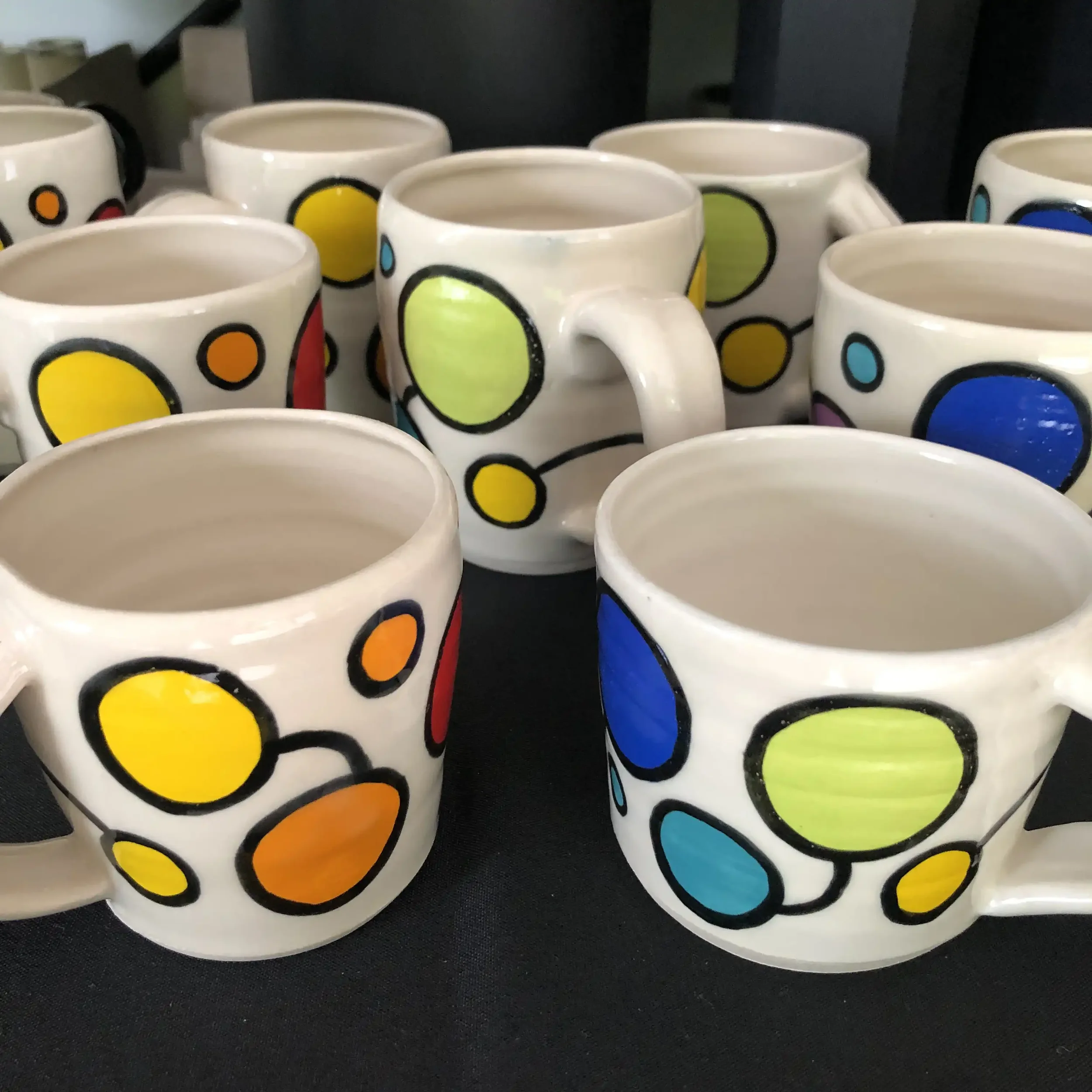 Colorful ceramic mugs with abstract dot pattern in yellow, orange, green, blue, purple, and red, arranged on a black surface.