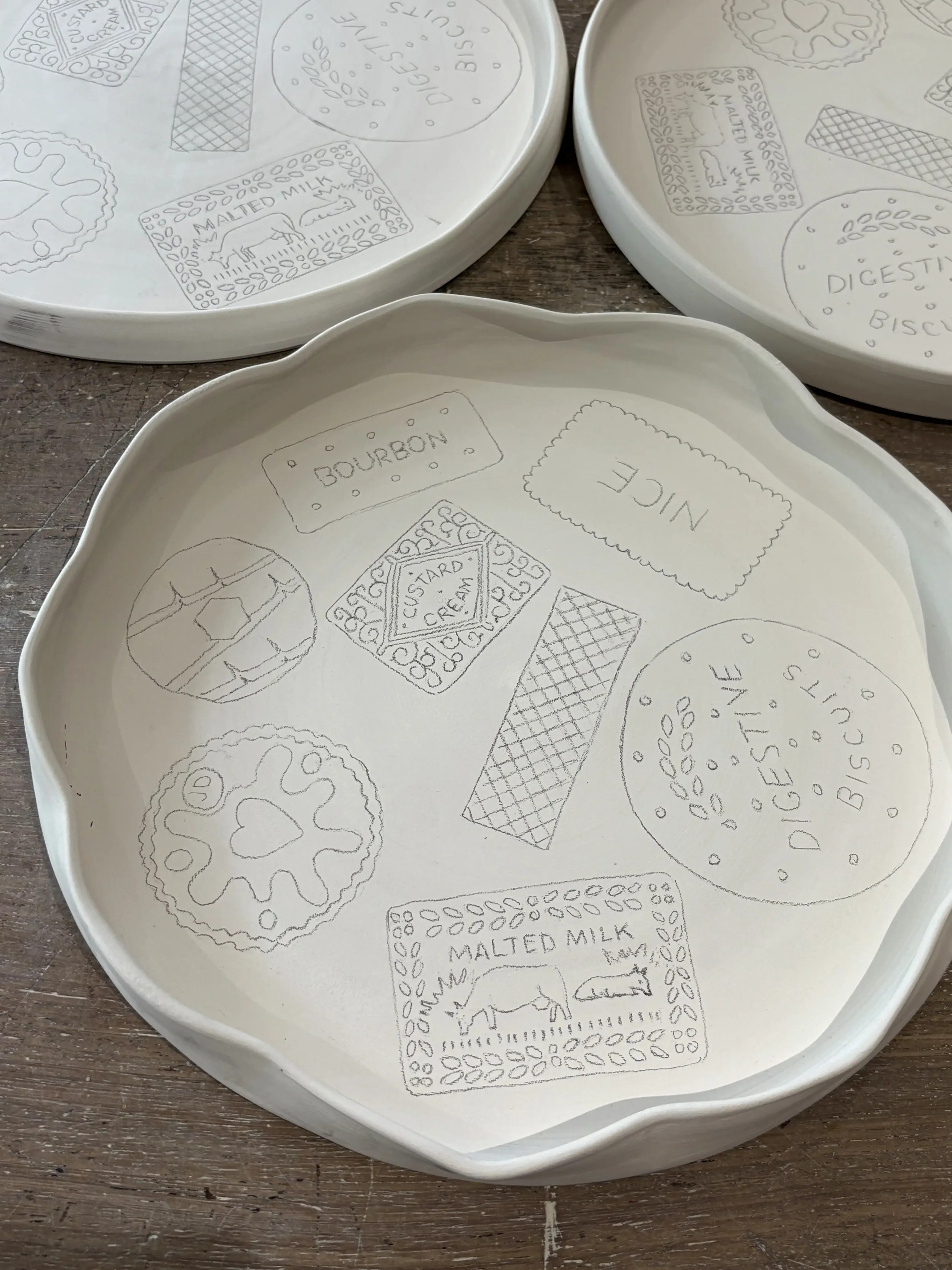 White ceramic plate with pencil sketches of various British biscuits, including a Custard Cream, Jammie Dodger, Rich Tea Biscuit and Digestive Biscuit. The plate rests on a wooden surface.