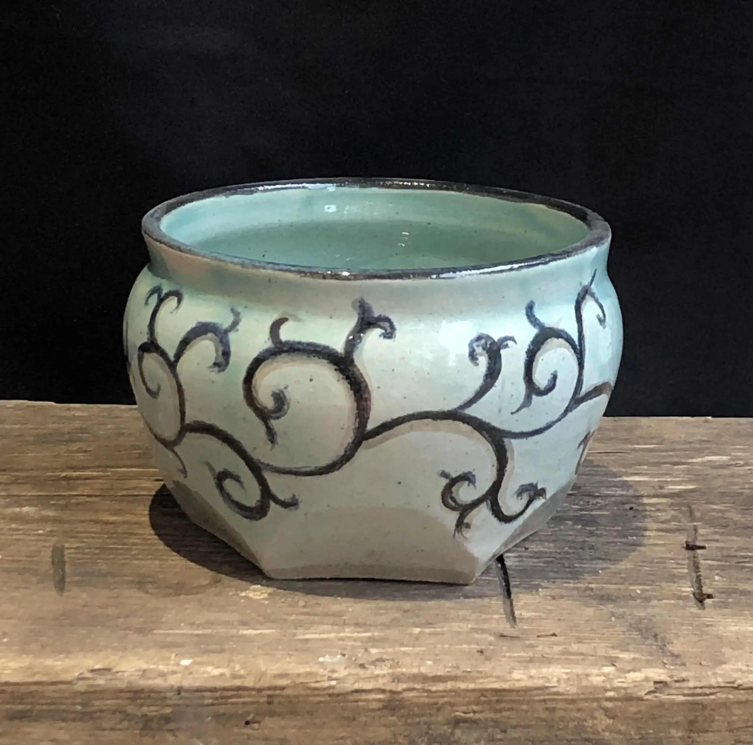 A turquoise ceramic bowl with a hand painted swirl design on a wooden surface, with a black background.