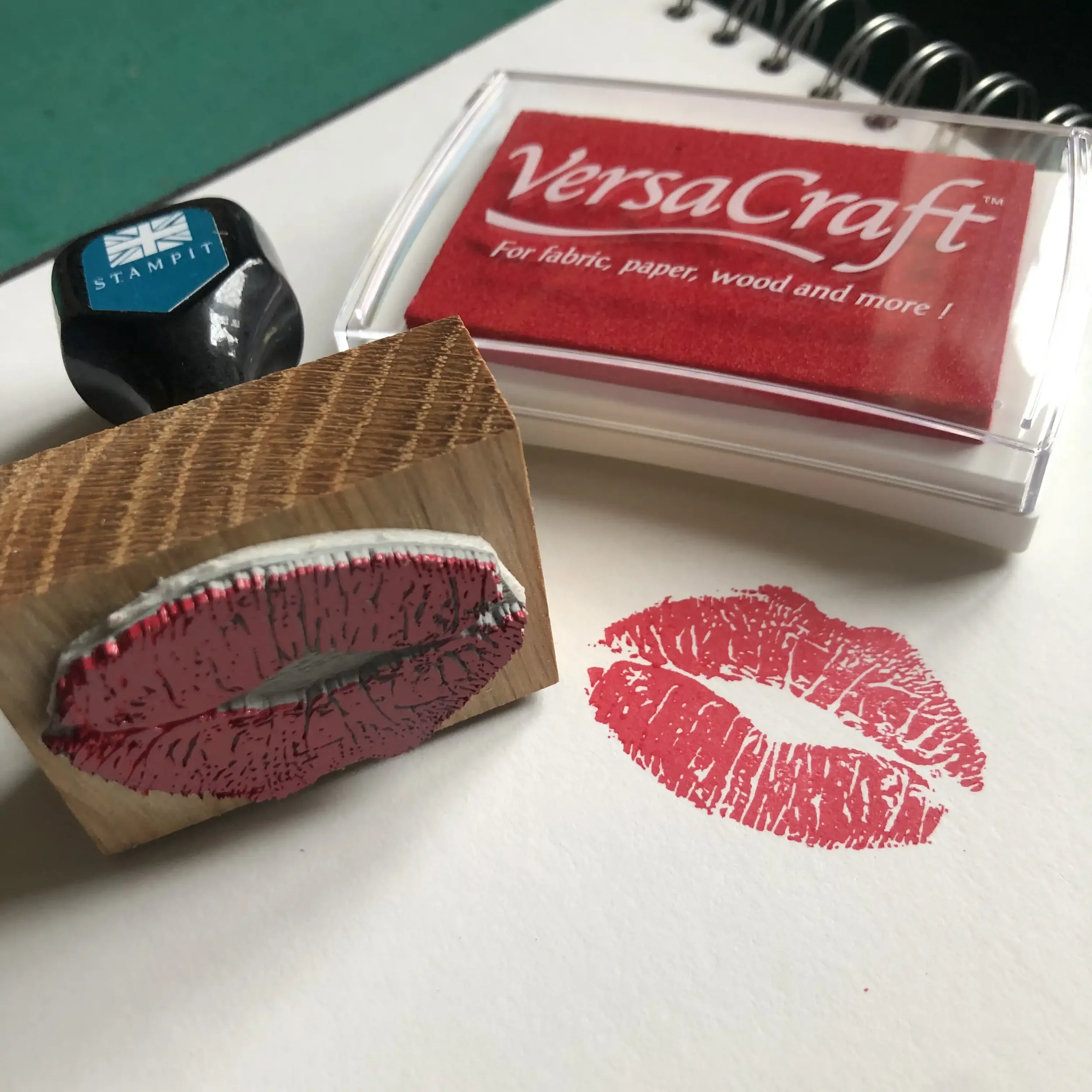 A rubber stamp with a lip print design, a red Versacraft ink pad for fabric, paper, and wood. The stamped lip print is on white paper.