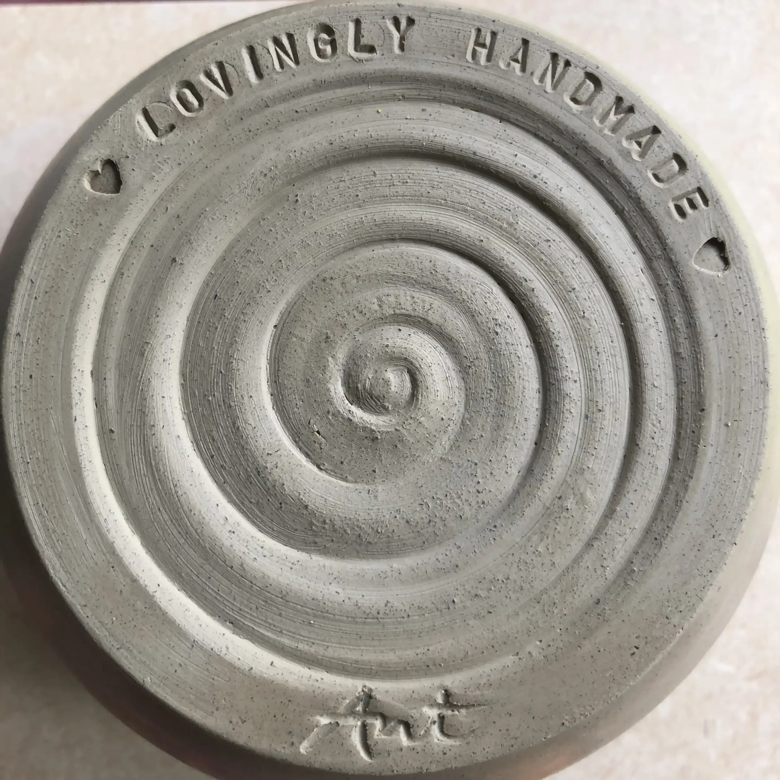 Close-up of the bottom of a clay bowl, stamped with the words 'LOVINGLY HANDMADE' along the edge, small hearts stamped at intervals.