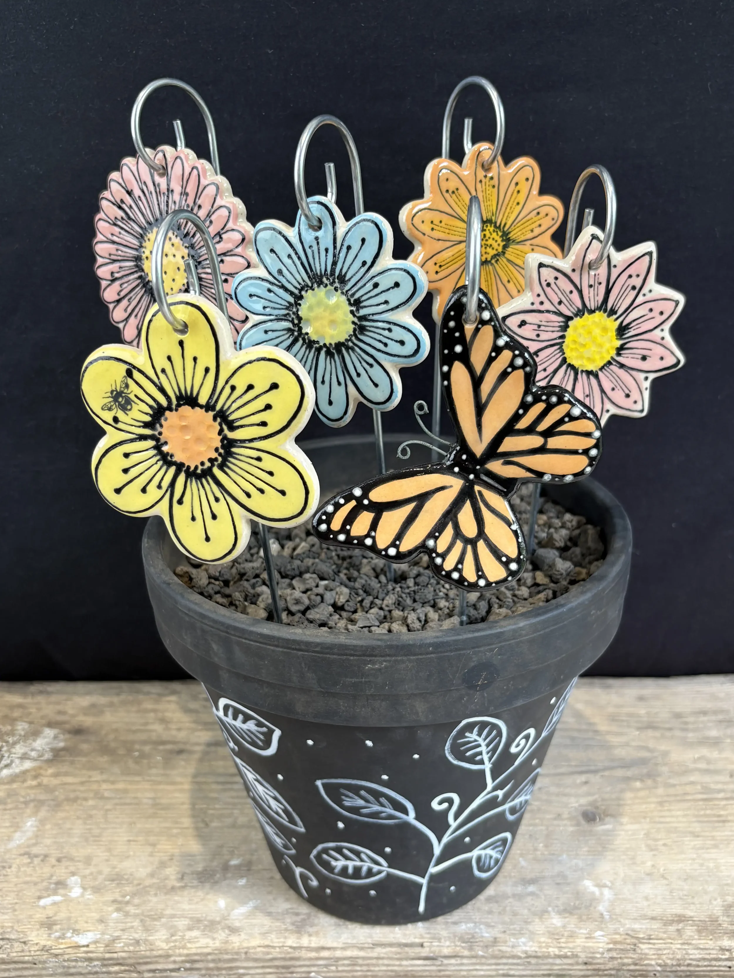 Colourful flower and butterfly decorations on metal stakes in a black pot with white painted leaf and swirl designs, set on a wooden surface against a black background.
