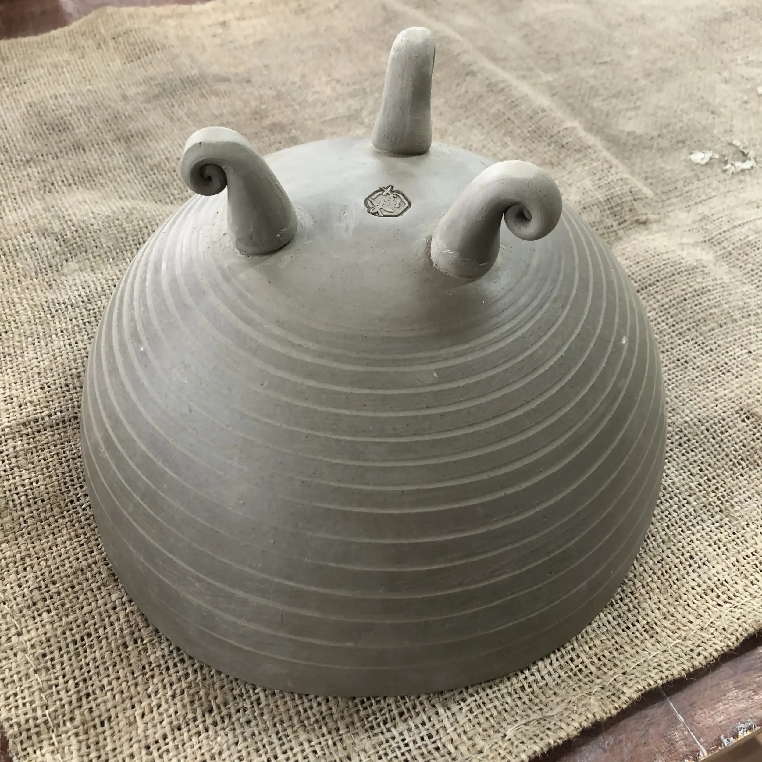 An upside-down ceramic bowl with tripod legs, placed on a hessian surface.
