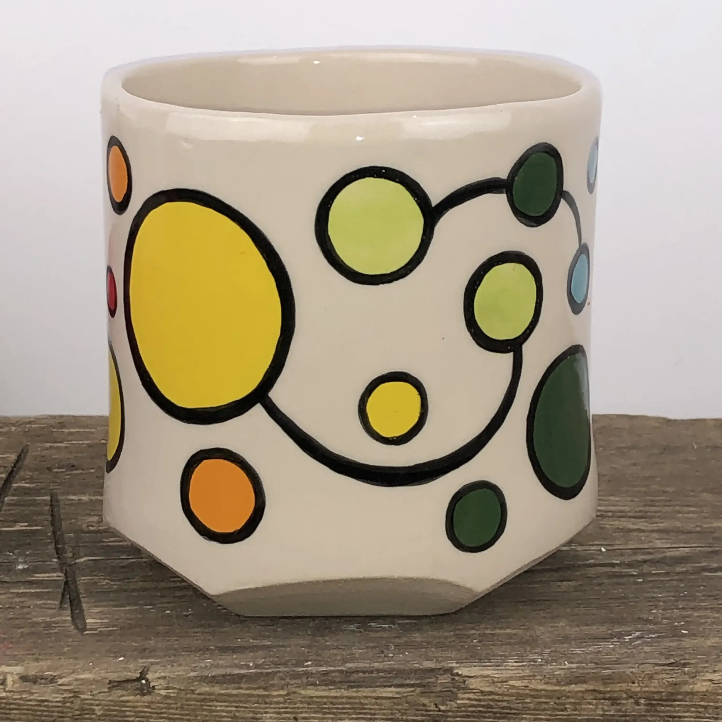 A ceramic planter with colourful circle patterns, featuring yellow, pale green, orange and dark green circles outlined in black, placed on a wooden surface.