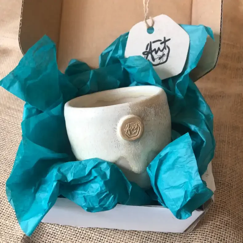 A cream glazed ceramic cup with an Ant Kelly Creations maker's mark, surrounded by turquoise tissue paper, inside a gift box with a tag.