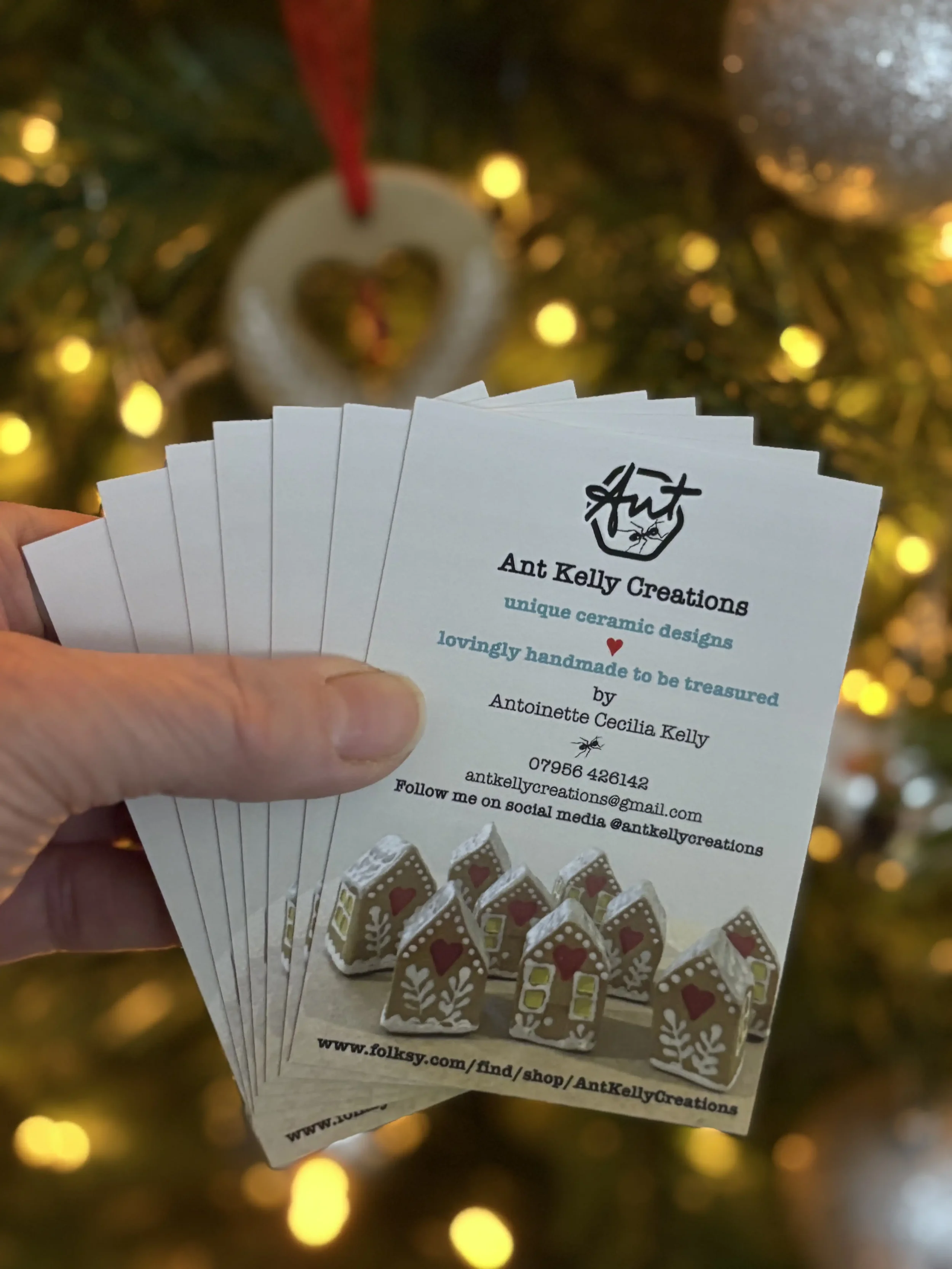 Business cards for Ant Kelly Creations featuring teeny ceramic gingerbread house ornaments, held in front of a decorated Christmas tree.
