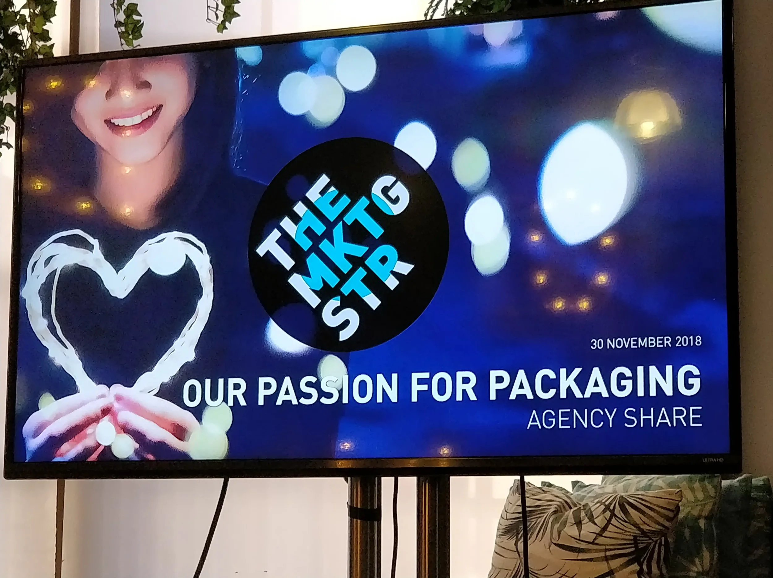 A woman smiling and holding a heart-shaped outline made of light, with a background of blue and white broken lights, and text on the screen that reads 'The Marketing Store' and 'Our Passion for Packaging, Agency Share', dated November 30, 2018.