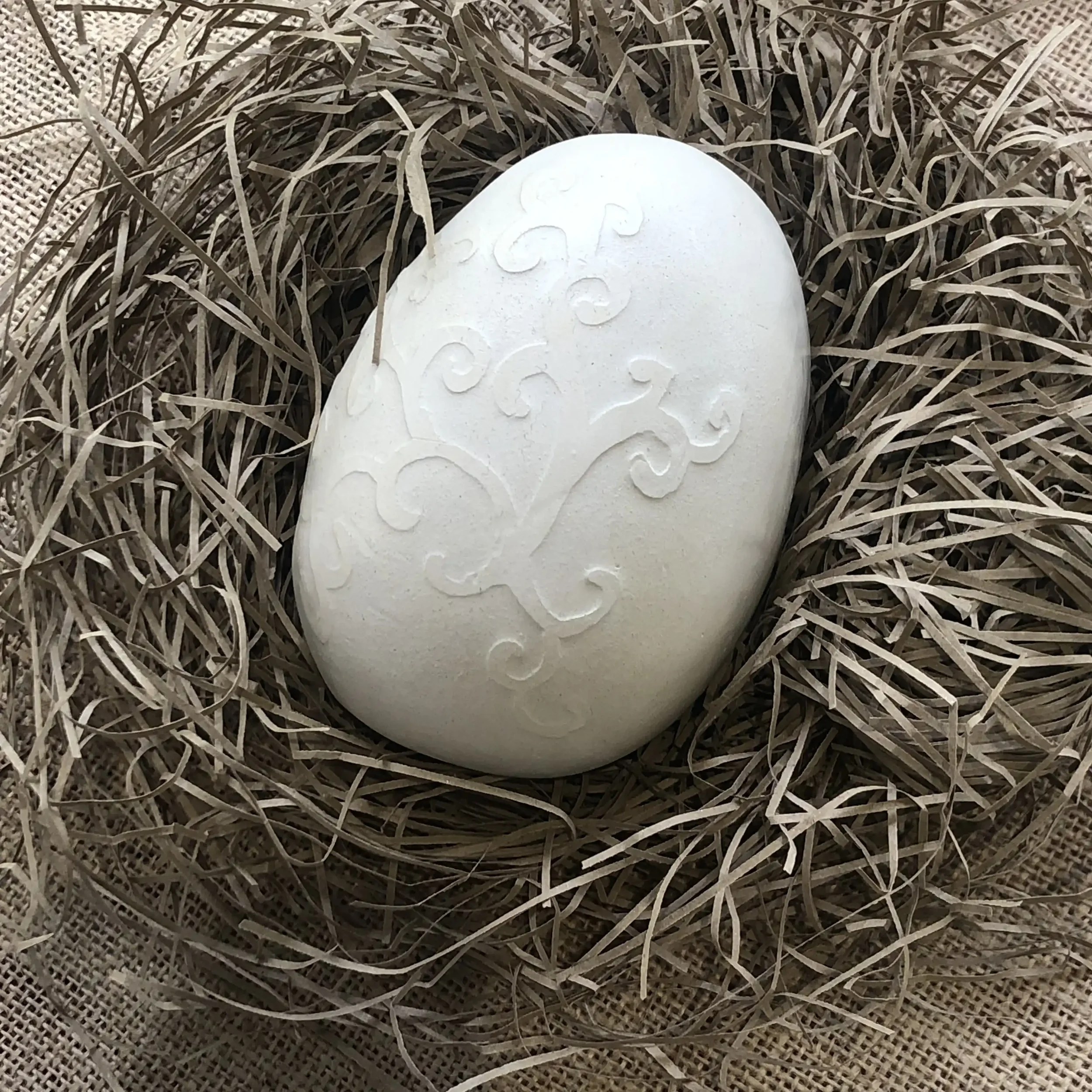 White decorative oval egg with an embossed swirl pattern, nestled in a bed of straw on a hessian surface.