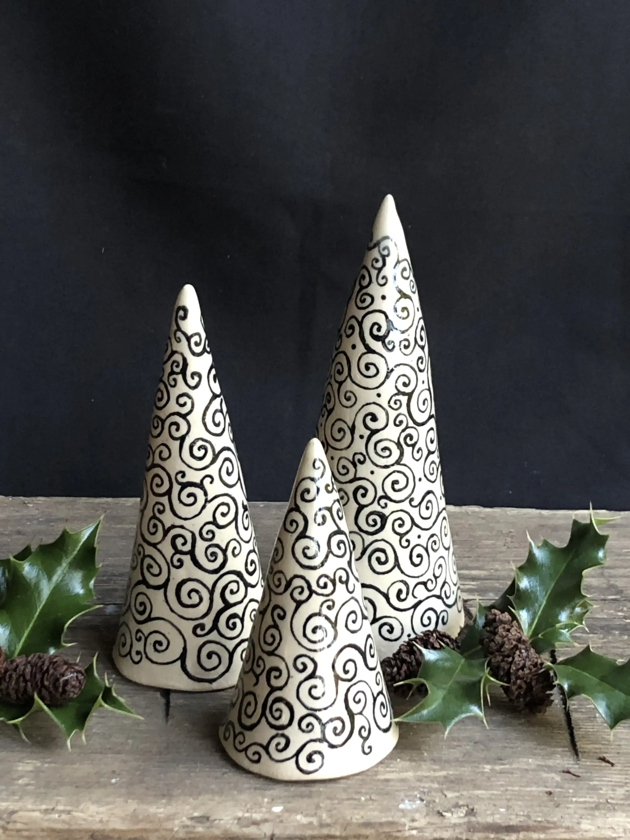 Three ceramic cone-shaped sculptures with black swirl patterns on a wooden surface, decorated with holly leaves and pinecones, against a dark background.