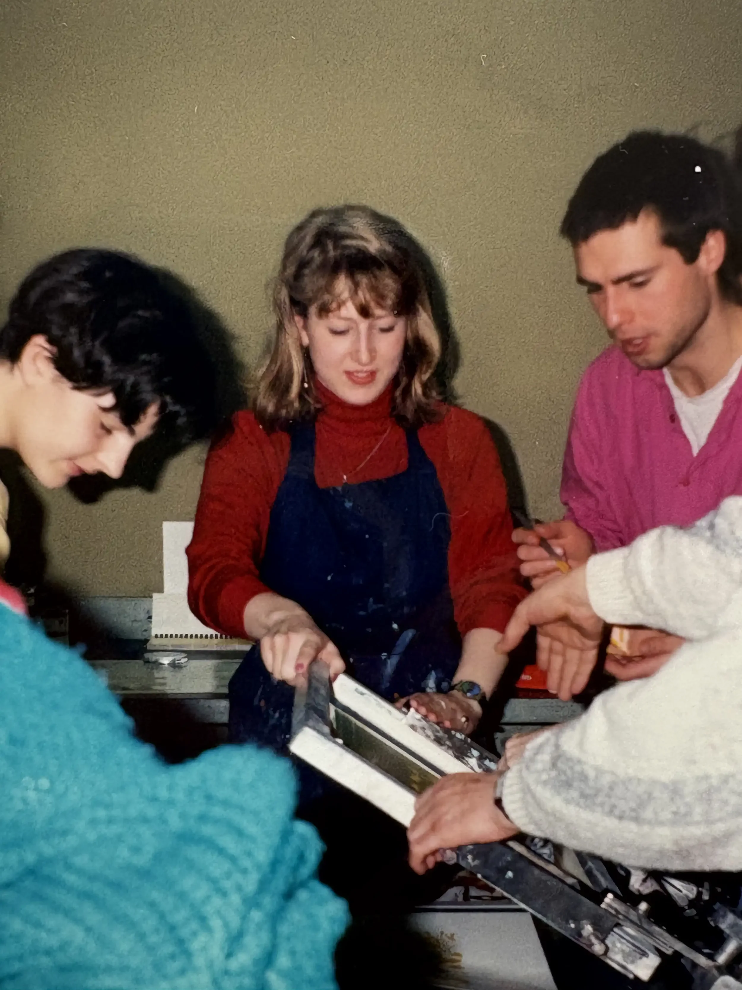 Four people gathered around a silkscreen bench, examining a framed item, with one person in a blue apron holding it.