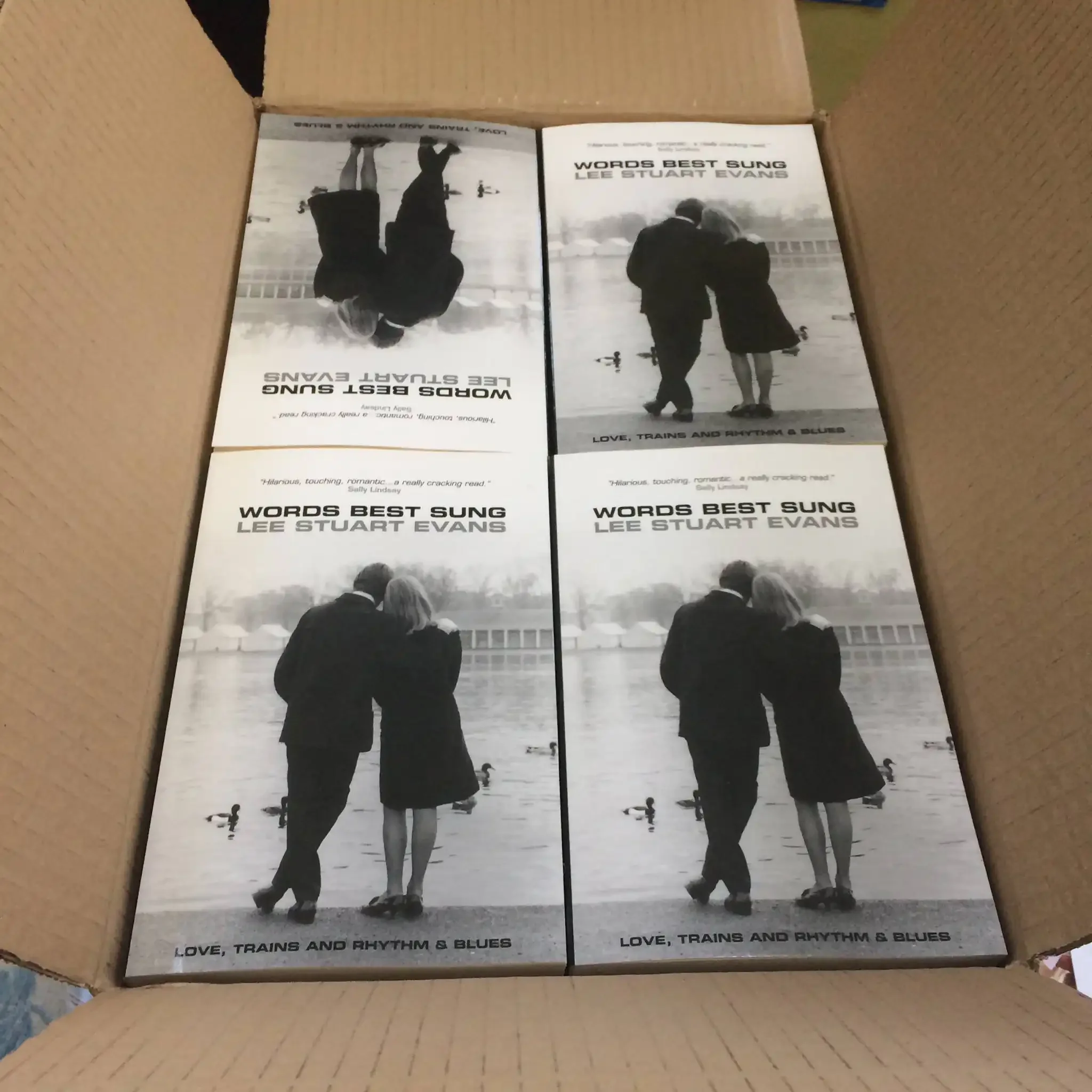 Box of four copies of the book "Words Best Sung" by Lee Stuart Evans, featuring a black and white cover image of an older couple walking by a river, with ducks and a bridge in the background.