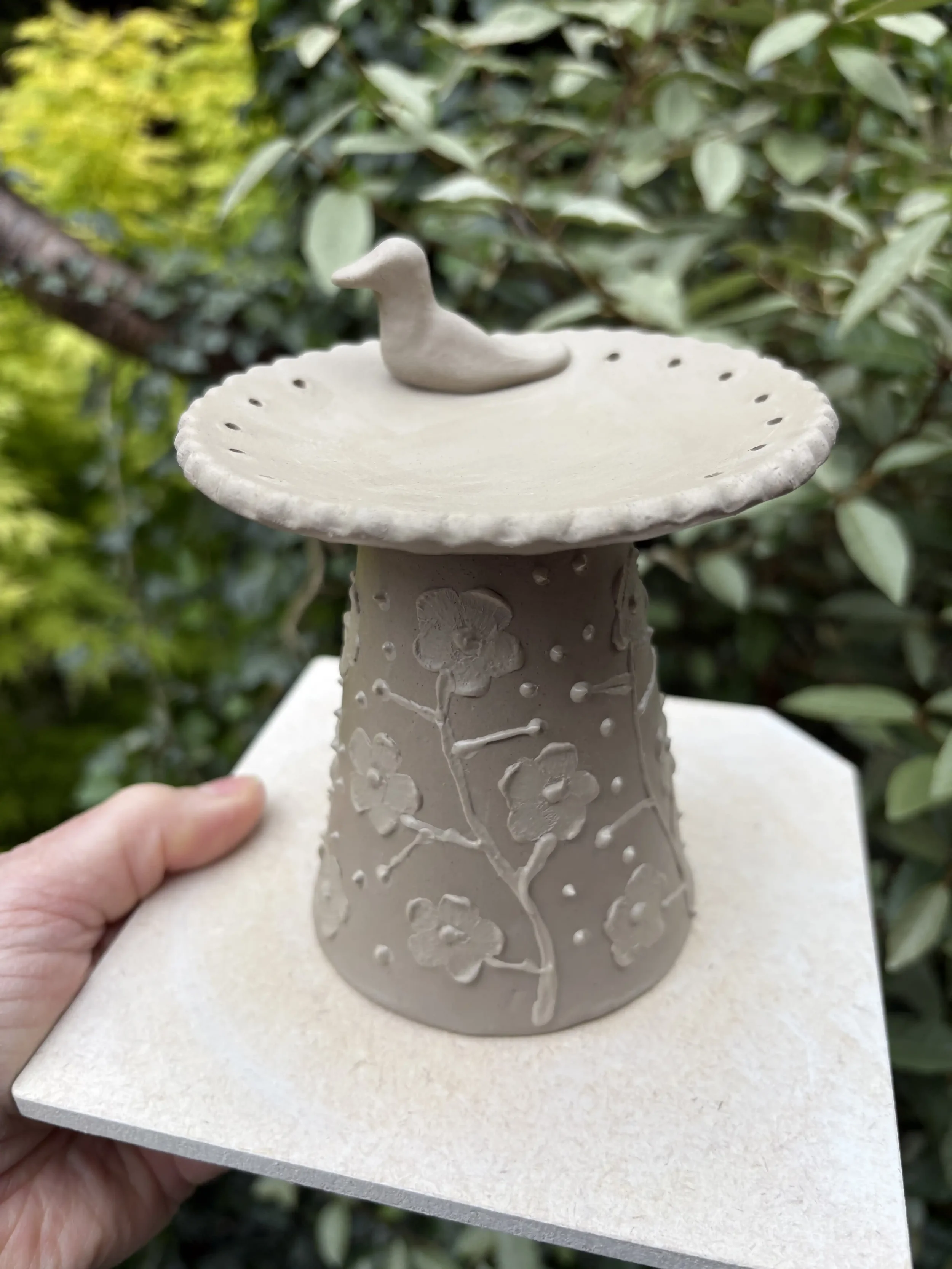 A ceramic jewellery stand in the form of a small birdbath with a base decorated with flower and branch patterns, topped with a shallow dish and a small bird figurine, held outdoors against green foliage.