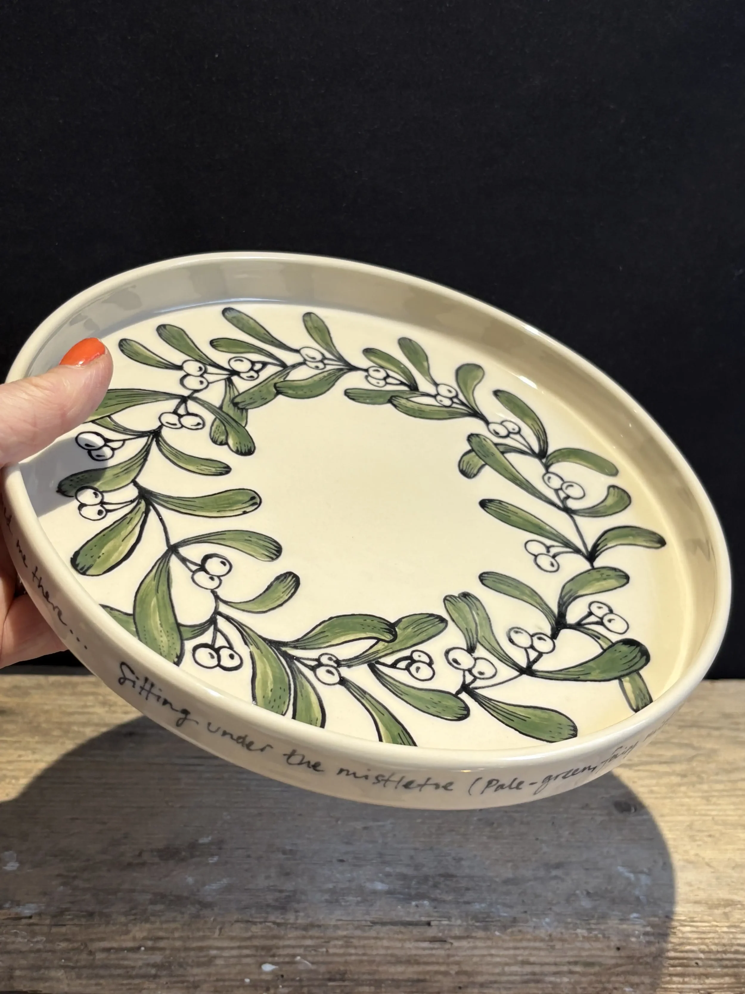 Decorative ceramic plate with green and white mistletoe design and a handwritten phrase around the edge, held by a person with painted nails against a black background and wooden surface.