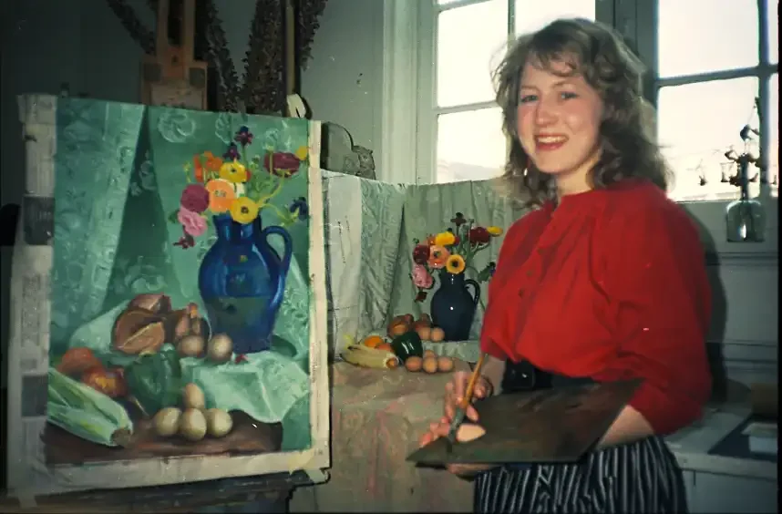 A young woman with curly hair wearing a red blouse and striped skirt, holding a palette and brush, painting a still life of a blue vase with colorful flowers, fruits, and vegetables on an easel in a well-lit room.