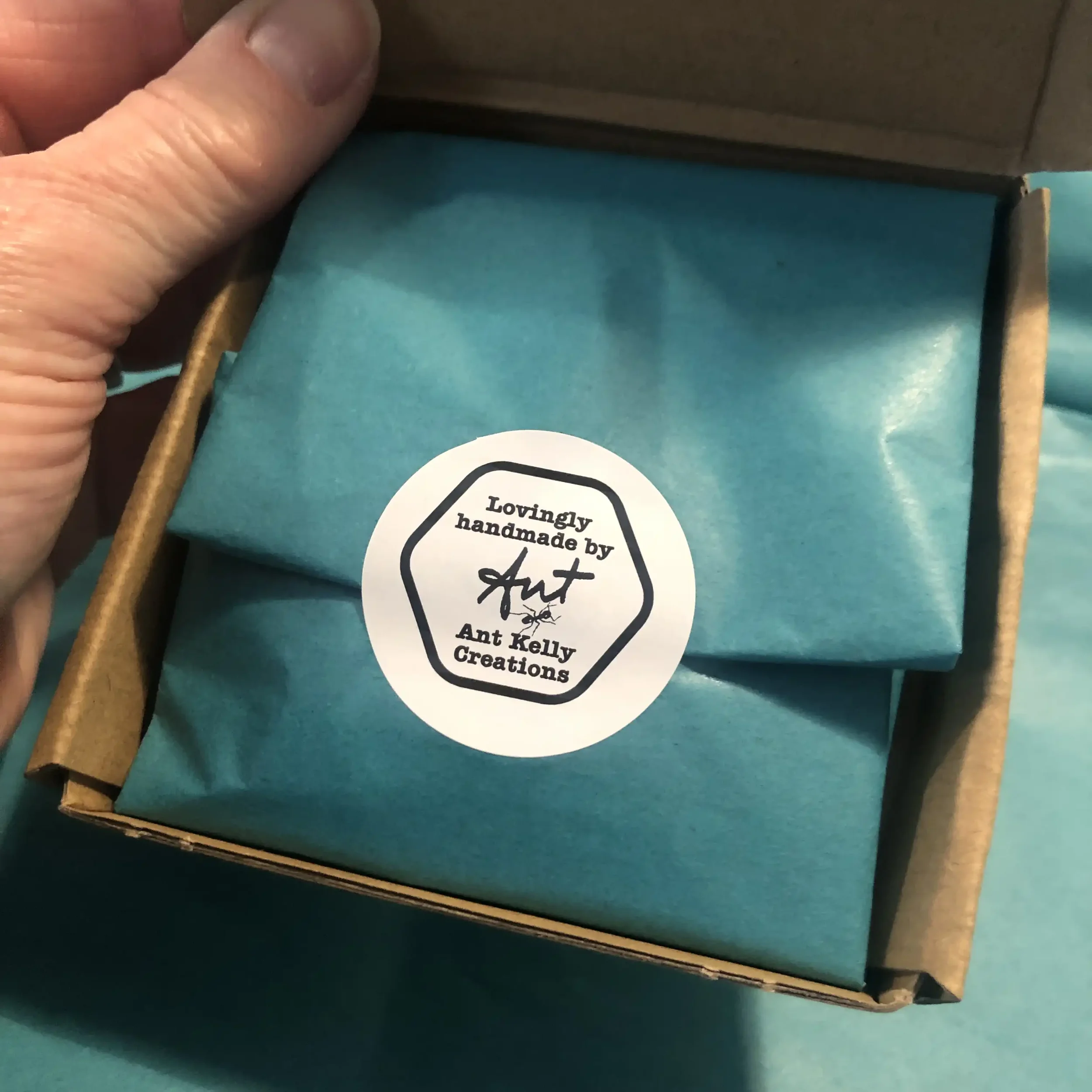 Open cardboard box containing wrapped items with turquoise tissue paper and a round sticker that says 'Lovingly handmade by Ant Kelly Creations'