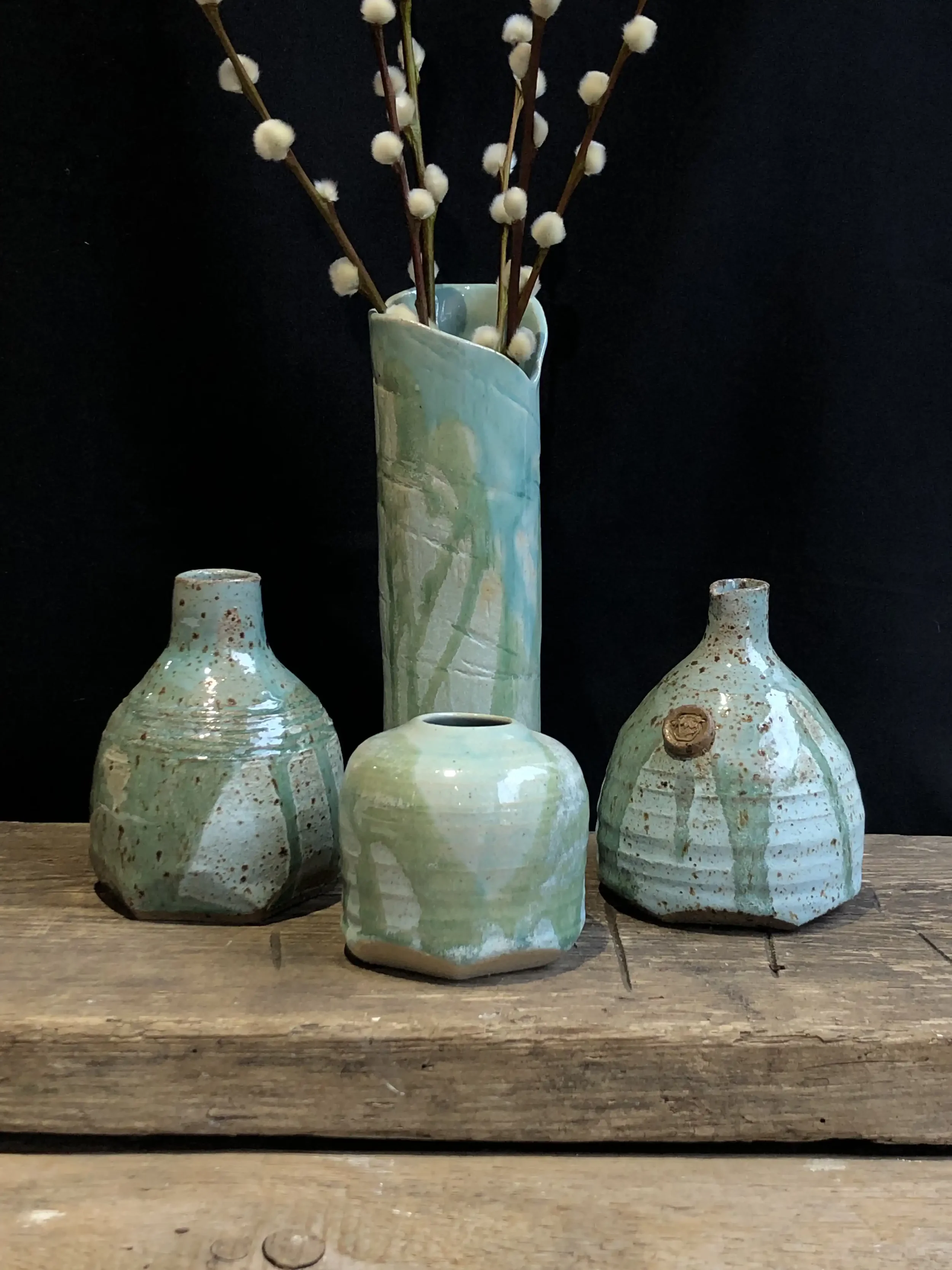 Set of four ceramic bud vases with greenish-glazed finish, displayed on a wooden surface against a black background; one tall vase in the back contains white fluffy pussy willow branches.