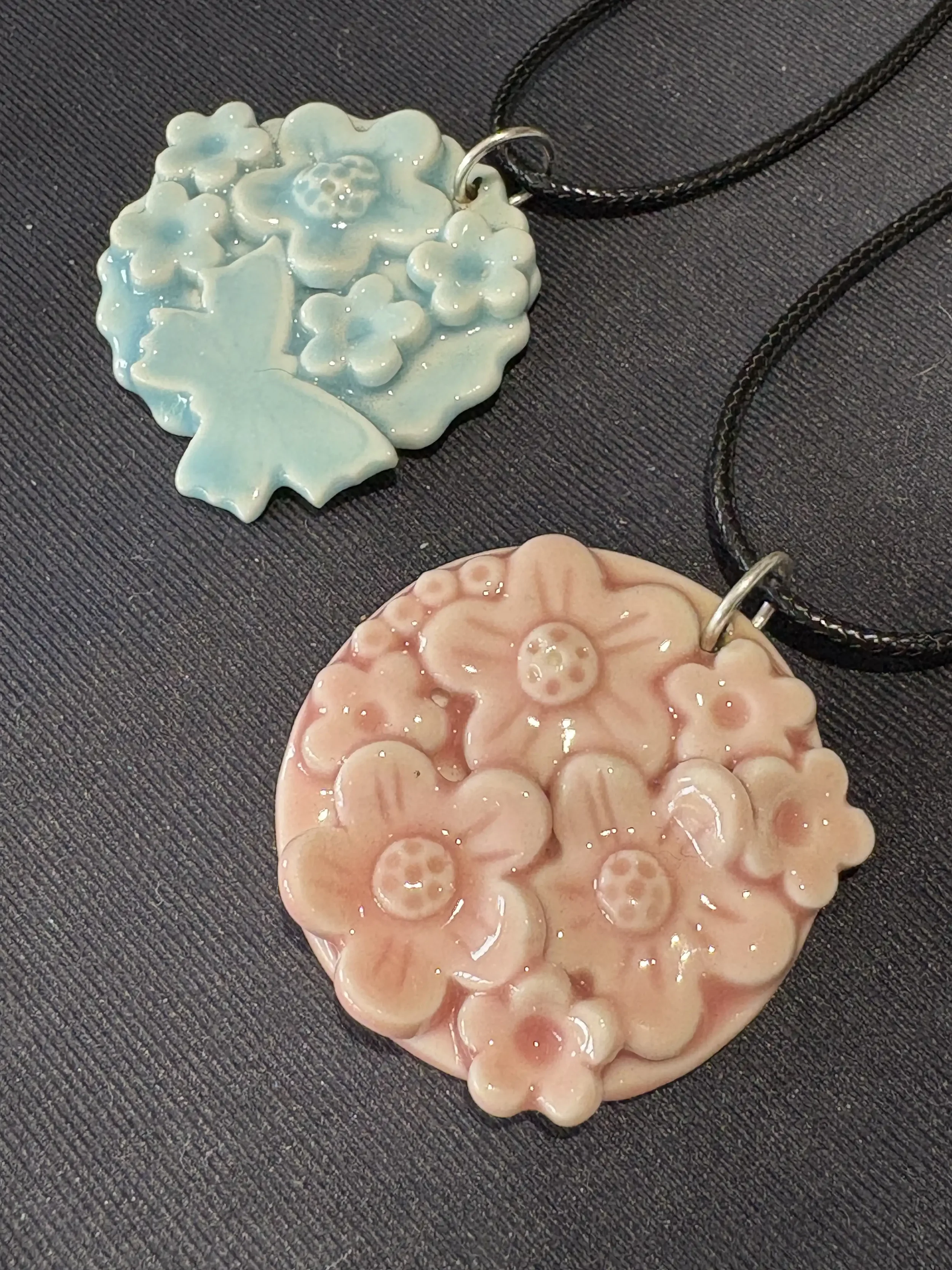 Close-up of two floral-shaped pendants, one blue and one pink, with shiny, textured surfaces, attached to black necklaces on a black textured background.