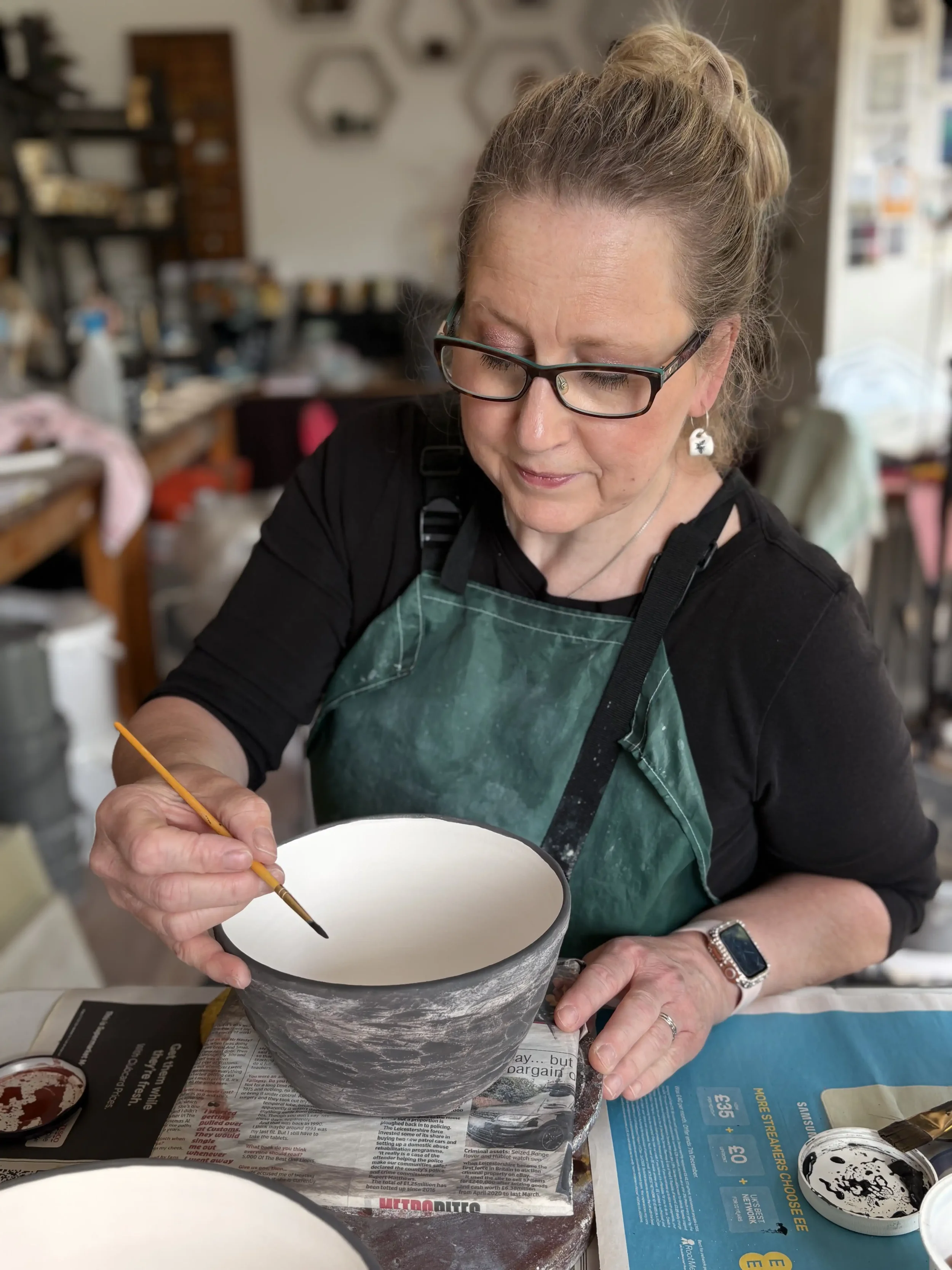 A woman with glasses and earrings is painting a ceramic bowl with a paintbrush in a pottery studio.