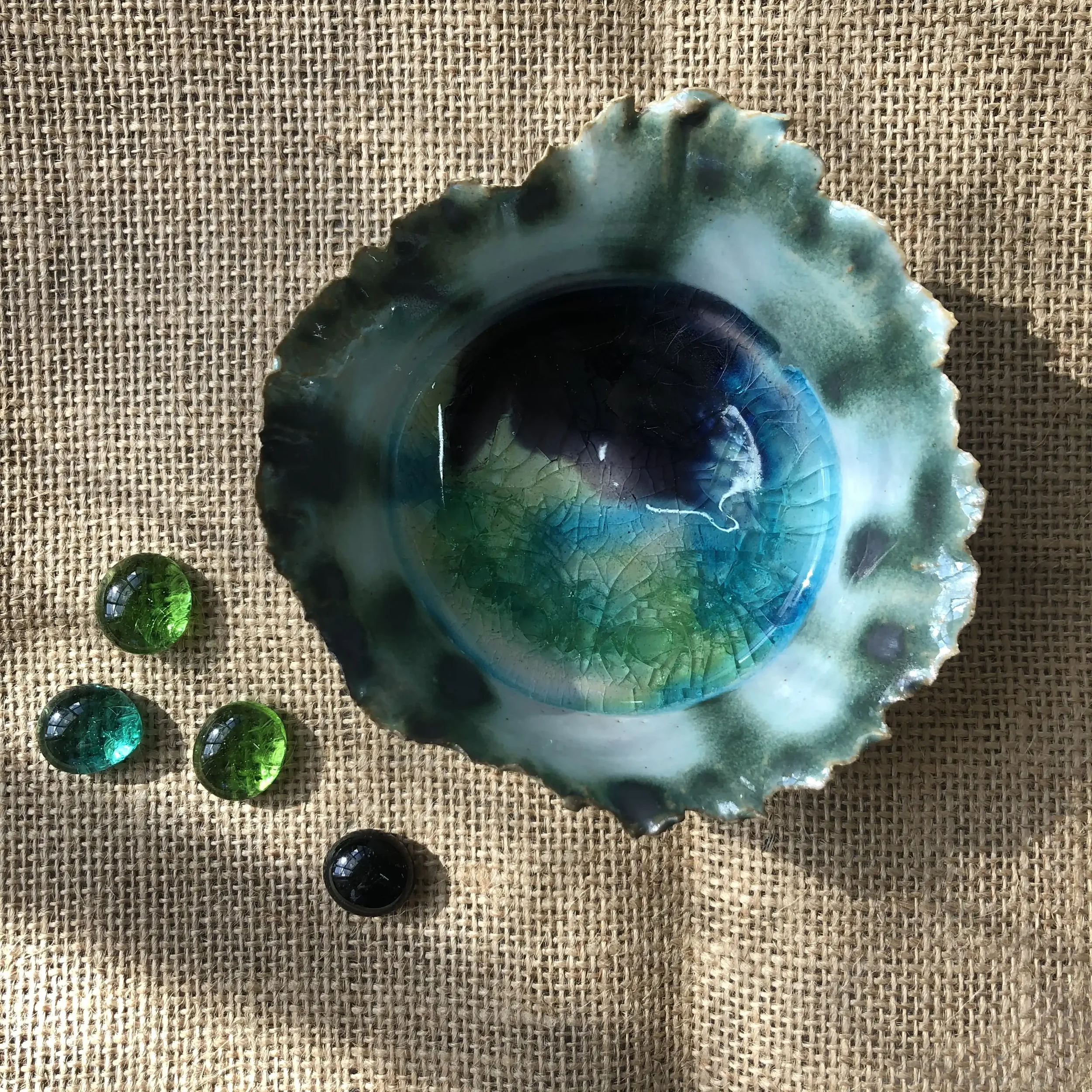 A small glazed dish with a ruffled edge, placed on a hessian surface. Four smaller, round glass stones in shades of green and black are positioned nearby.