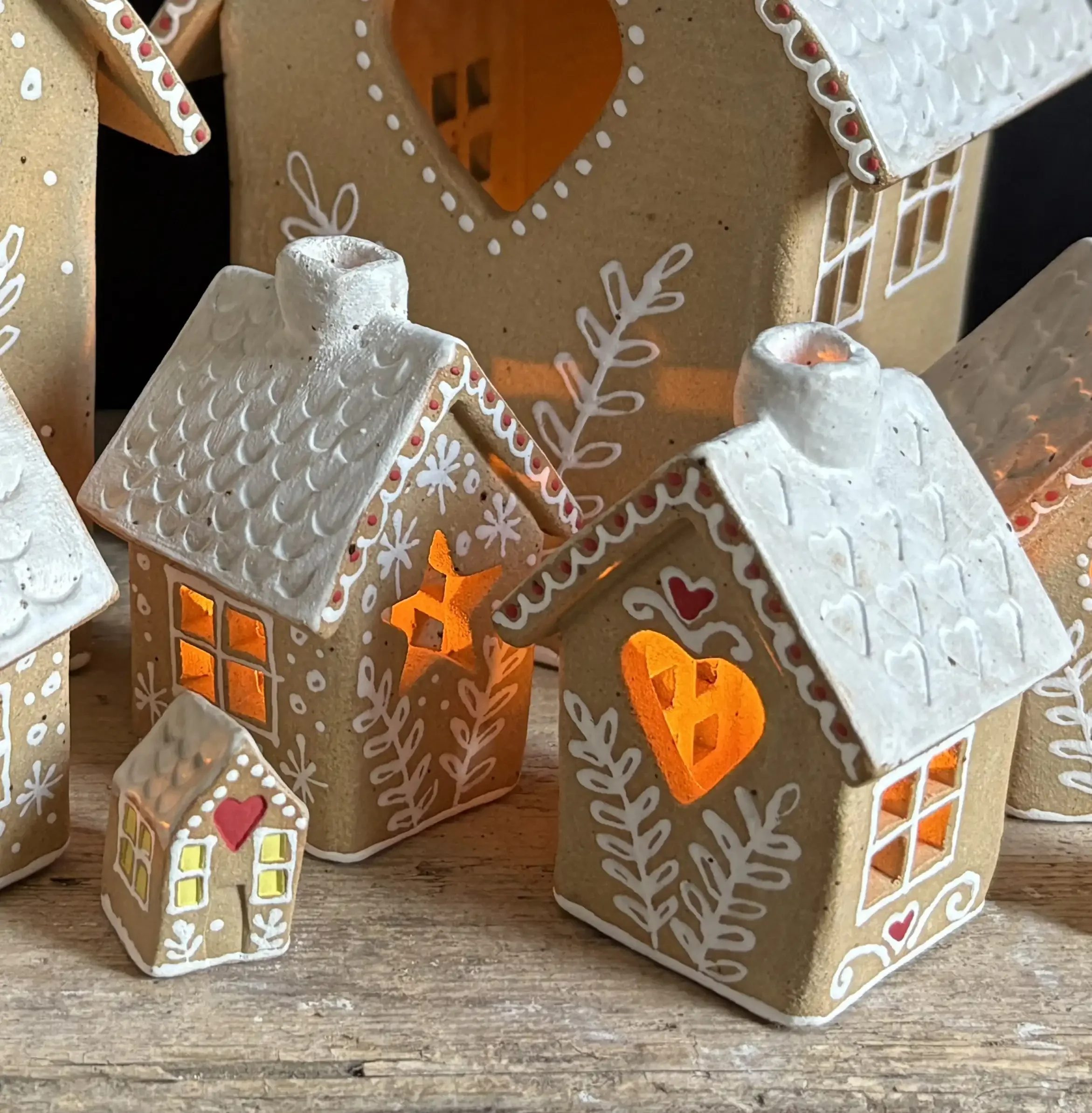 Multiple gingerbread-style ceramic houses with illuminated windows, decorated with white icing-like designs and hearts, set on a wooden surface.