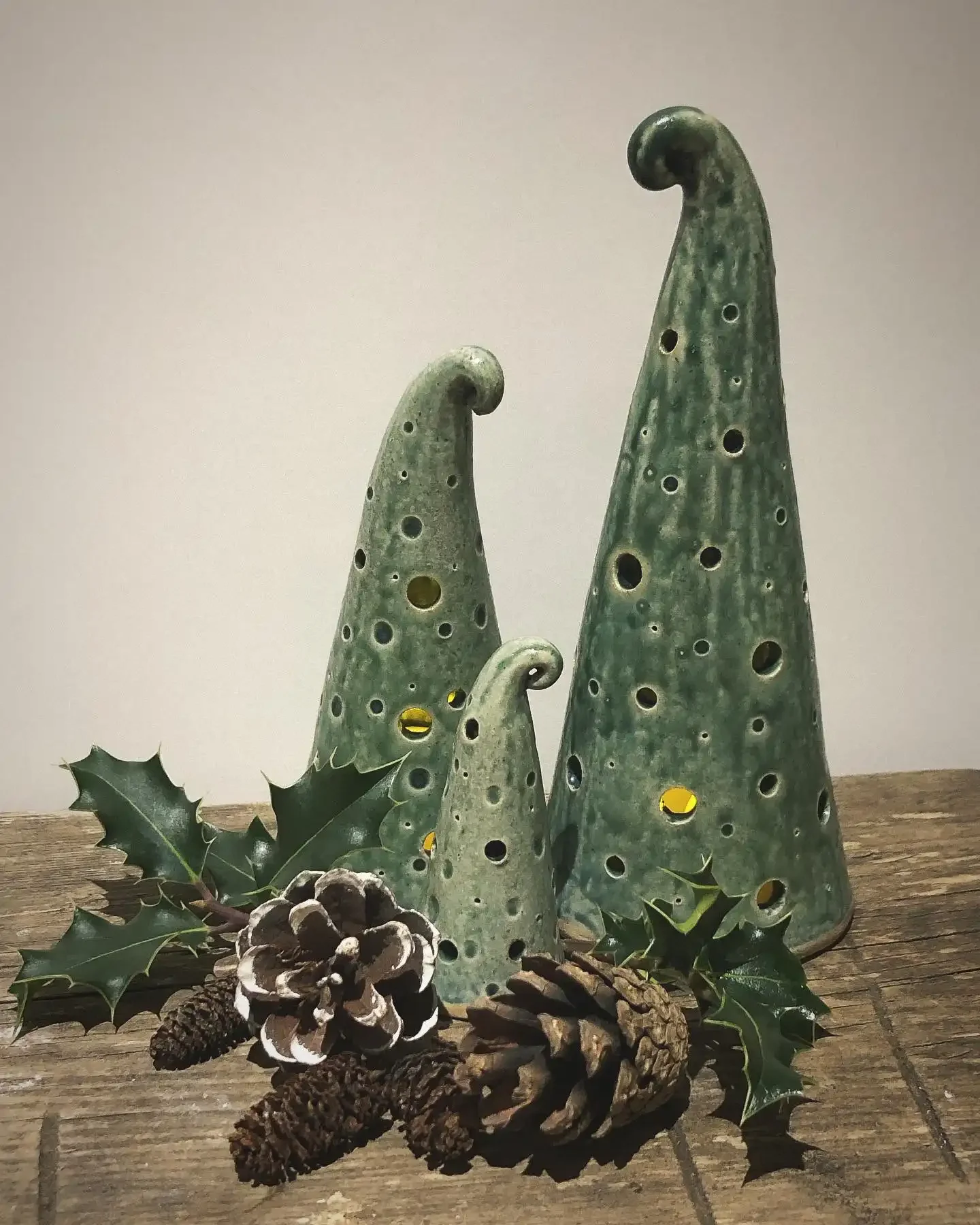 Three green ceramic Christmas trees with holes, surrounded by pinecones and holly leaves on a wooden surface.